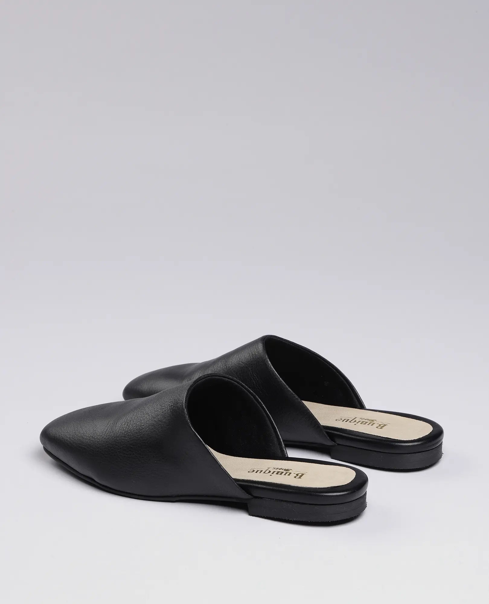 זוג כפכפים שטוחים-רוקס של B.unique מעור שחור עם עקב נמוך, מוצגים על רקע אפור בהיר.