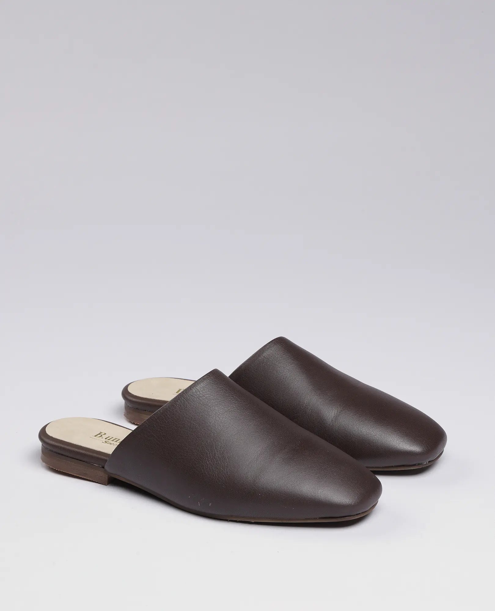 זוג כפכפים שטוחים מדגם "רוקס" של המותג B.unique בצבע חום, המוצגים על רקע אפור בהיר חלק.