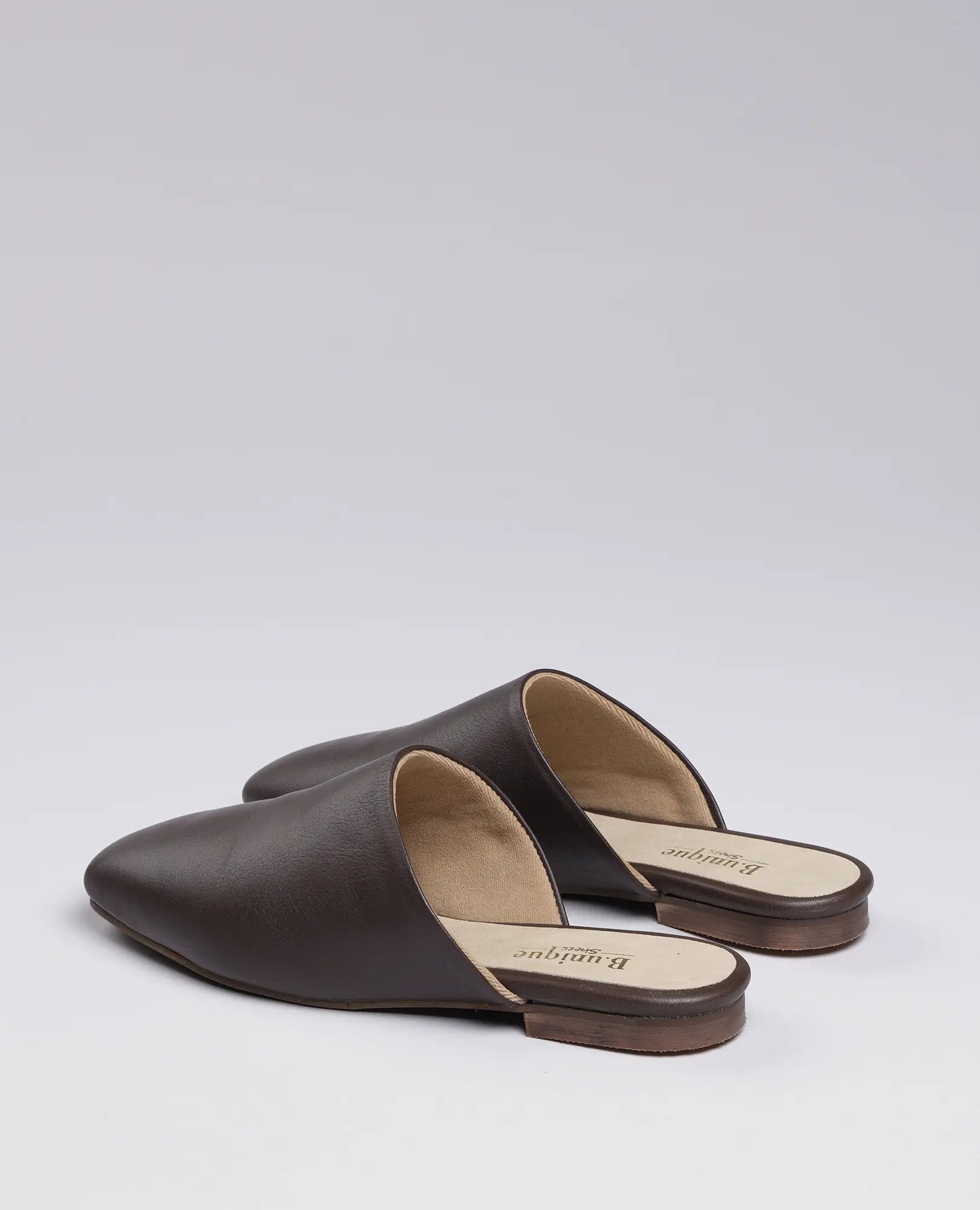 B.unique כפכפים שטוחים - רוקס: זוג כפכפים חומים מעור בעיצוב סגור עם סוליה שטוחה מעץ על רקע בהיר.