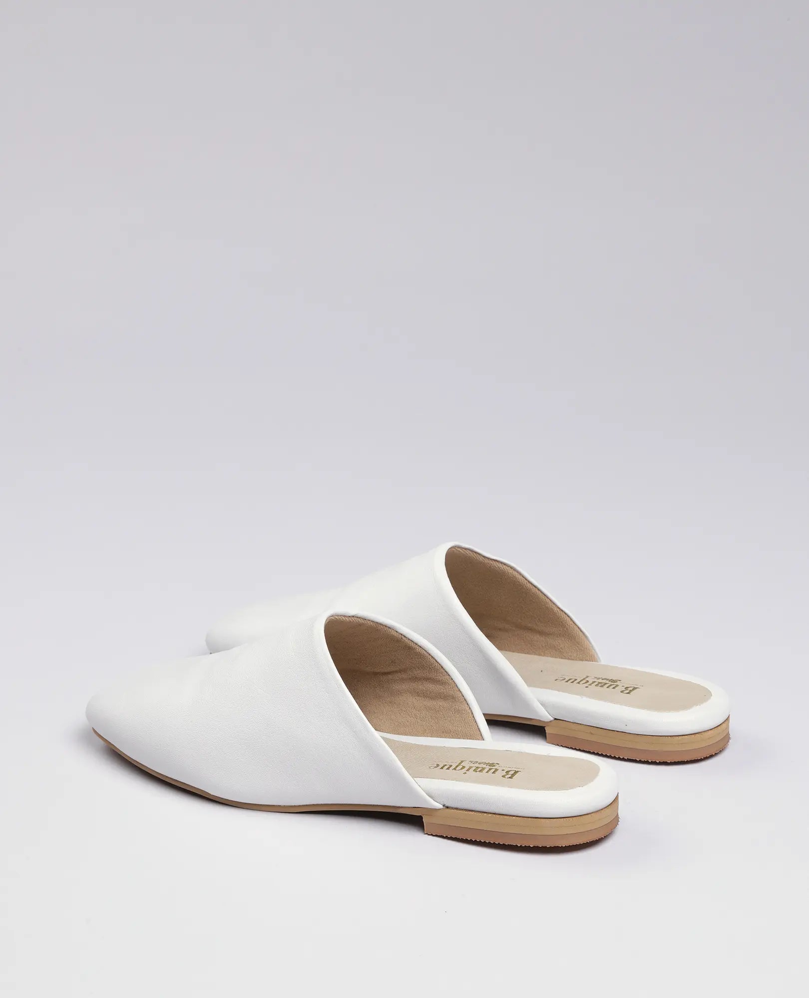 זוג כפכפים שטוחים של B.unique – נעלי מולה מעור לבן לנשים, עם סוליות שטוחות בצבע חום על רקע בהיר.