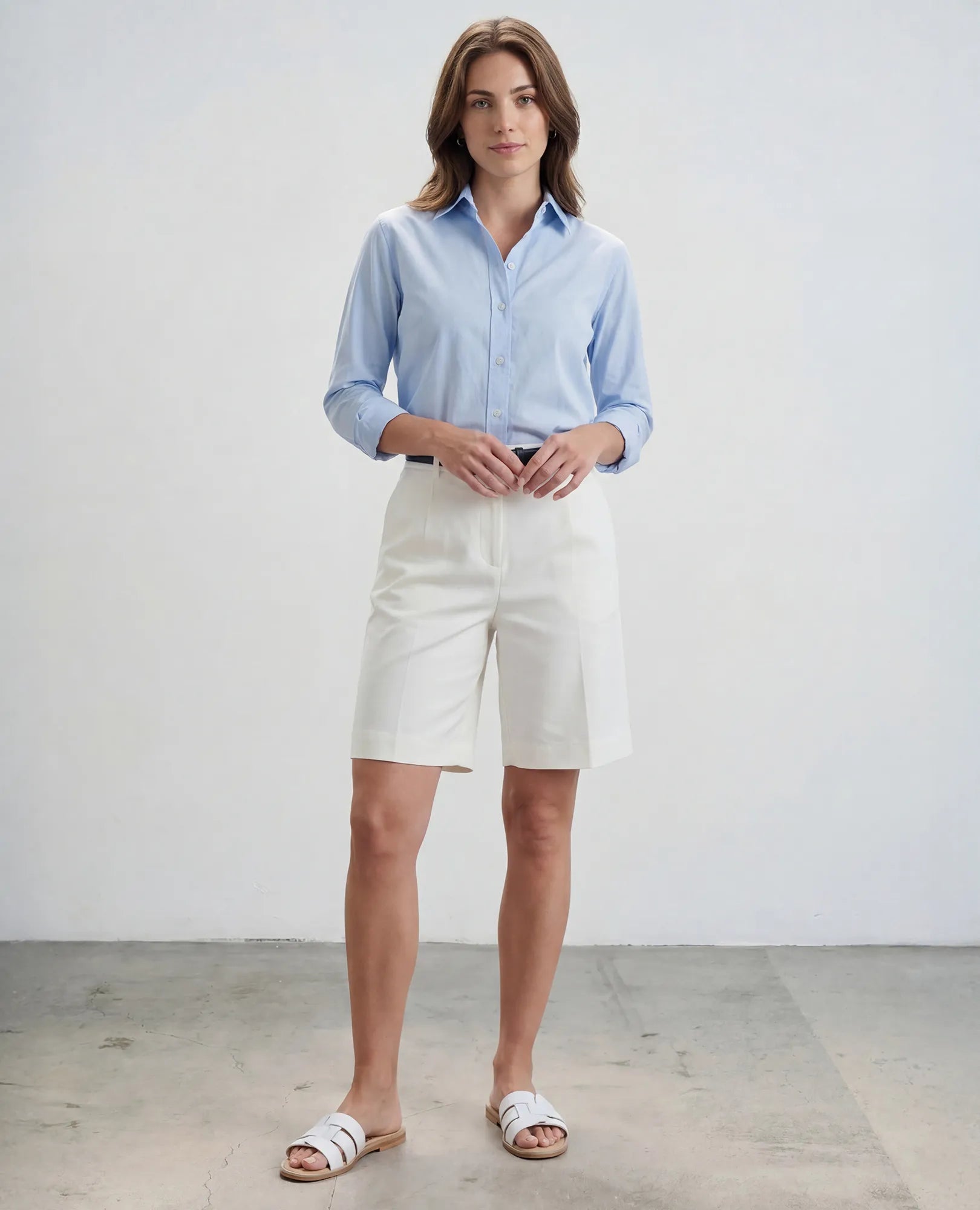 אישה לבושה בחולצה מכופתרת בצבע תכלת, מכנסיים קצרים לבנים וכפכפי רצועות-סקאיי של B.unique עומדת בחדר מינימליסטי.