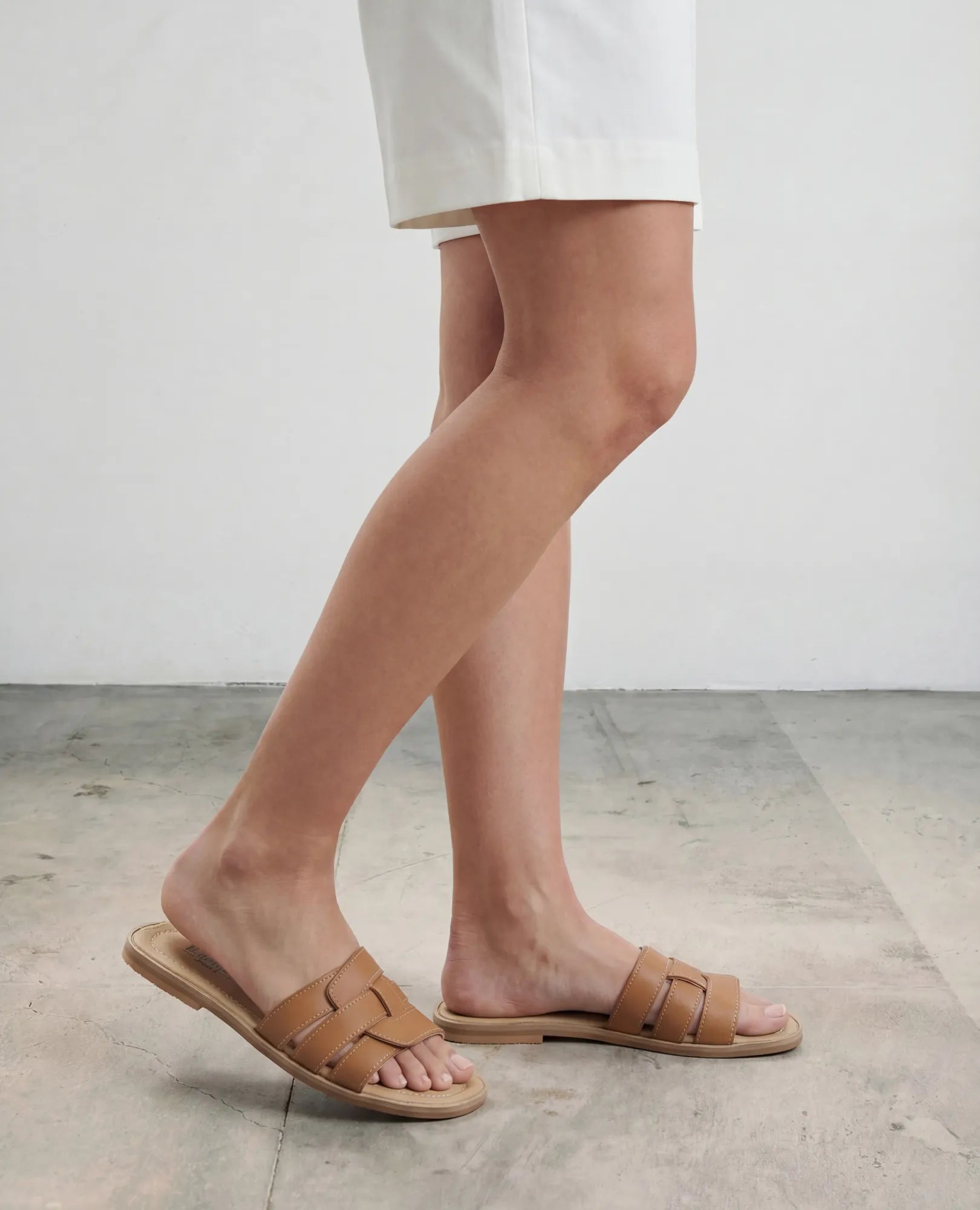 אדם לבוש במכנסיים קצרים לבנים וכפכפי רצועות של B.unique - סקאיי, עומד על רצפת בטון בהירה על רקע אחיד.