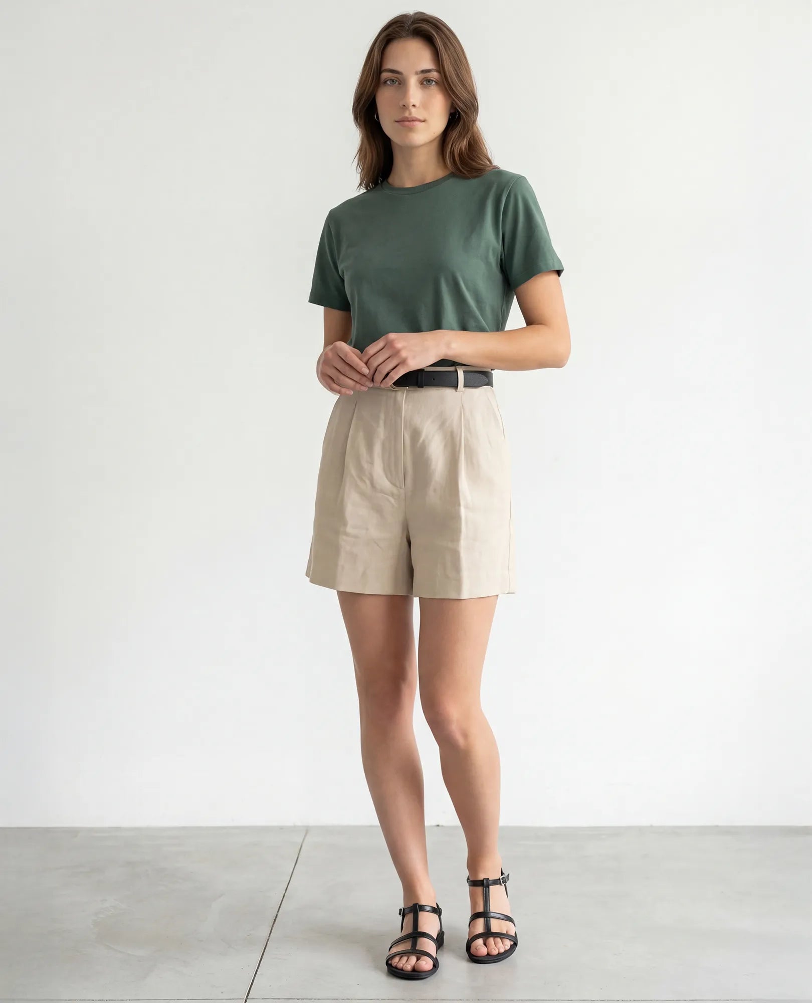 אישה בחדר מינימליסטי לובשת חולצת טי ירוקה, מכנסיים קצרים בצבע בז' וסנדלי גלדיאטור של B.unique – דגם קיילי.