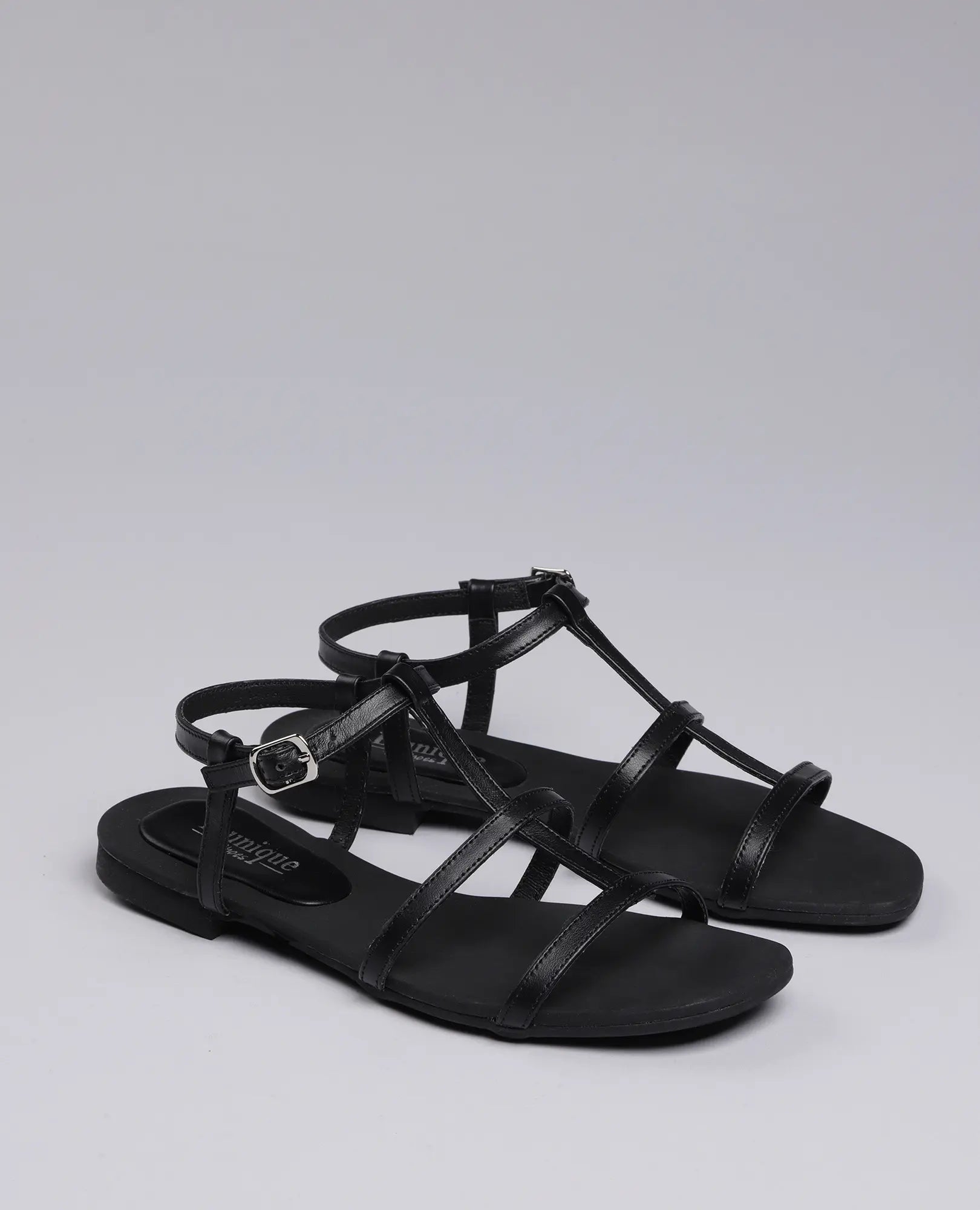 B.unique סנדלי גלדיאטור-קיילי: נעלי נוחות שטוחות בצבע שחור עם רצועות, אבזם קטן ועיצוב על רקע אפור בהיר.