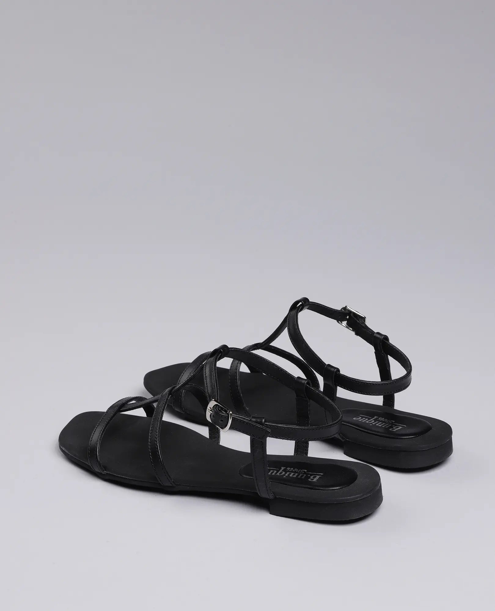 זוג סנדלי גלדיאטור-קיילי שחורים של B.unique, עם רצועות בקרסול ואבזמים, על רקע אפור אחיד.