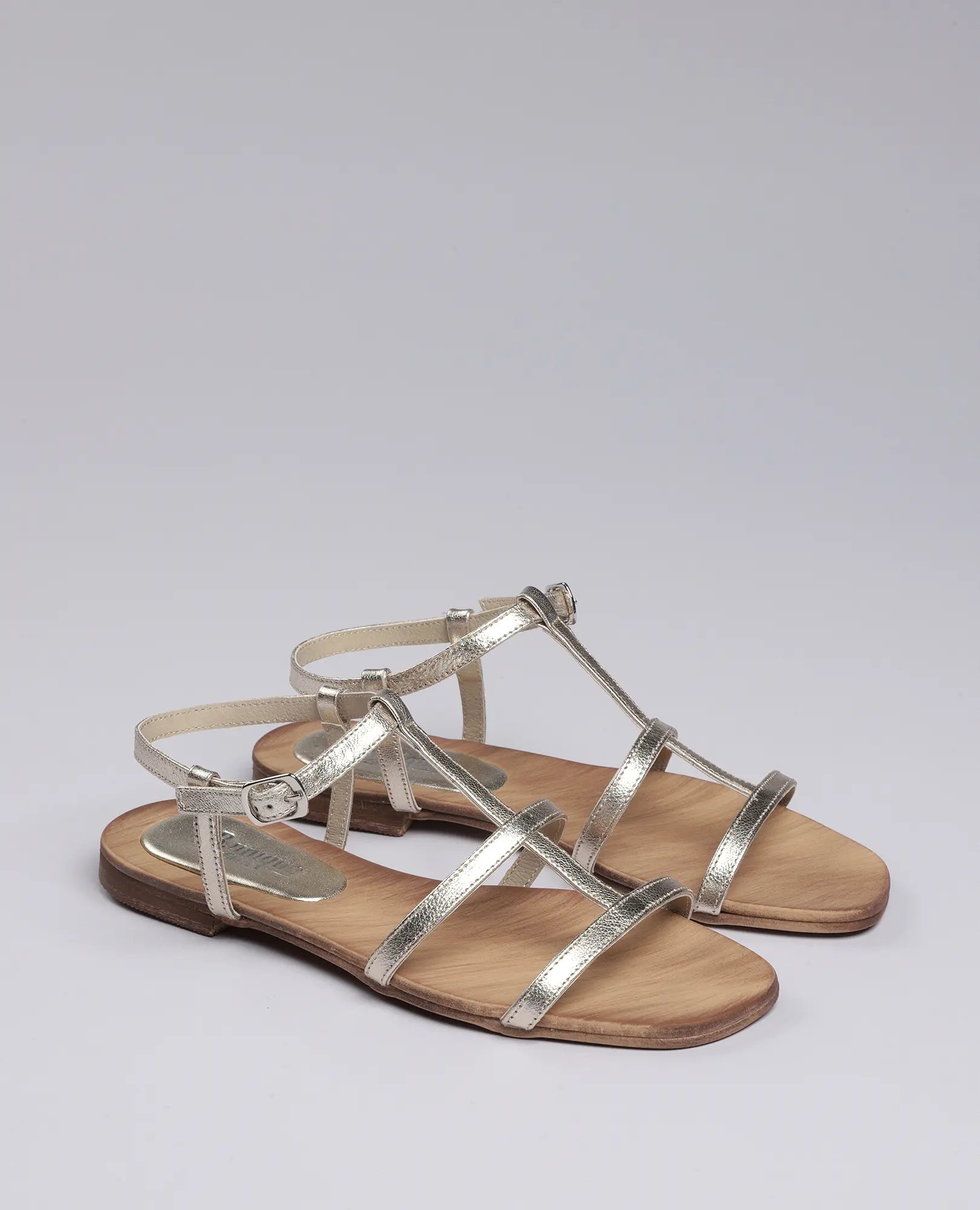 זוג סנדלי גלדיאטור – סנדלי רצועות שטוחות בצבע זהב מטאלי עם רצועות בקרסול מבית B.unique על רקע אחיד.