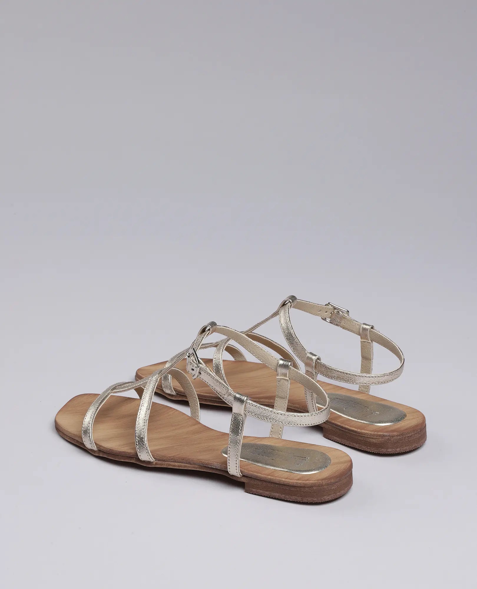 זוג סנדלי גלדיאטור "קיילי" מבית B.unique עם רצועות מתכתיות וסוליות עץ שטוחות, על רקע אפור בהיר.