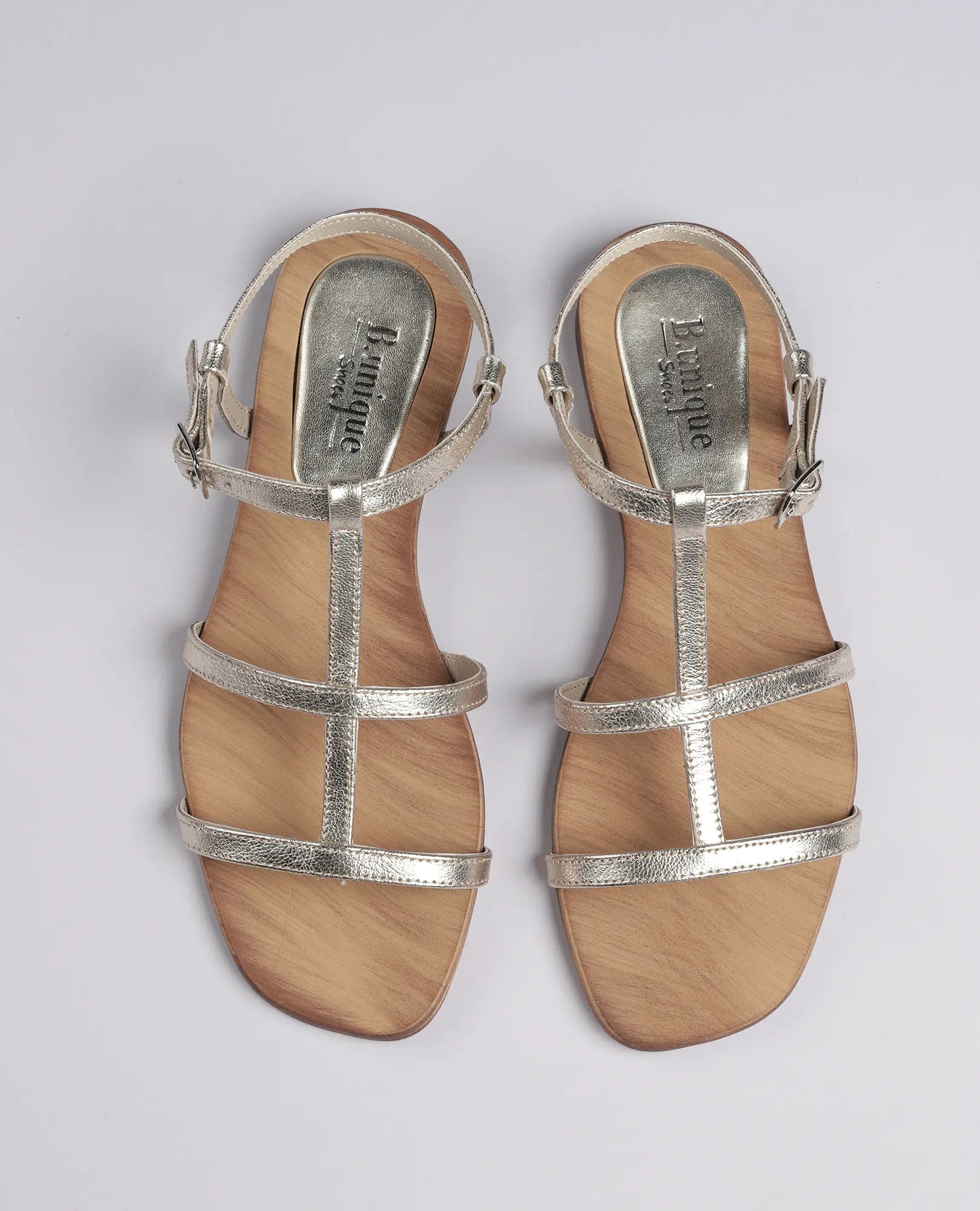 זוג סנדלי גלדיאטור-קיילי של B.unique בצבע כסף, עם רצועות ואבזמים בקרסול, על רקע בהיר.