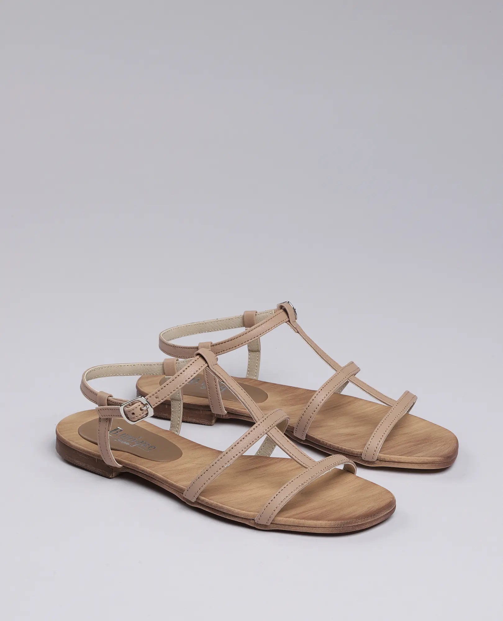 B.unique סנדלי גלדיאטור-קיילי: זוג נעליים שטוחות בצבע בז' עם רצועות ואבזמים, מוצגות על רקע בהיר.