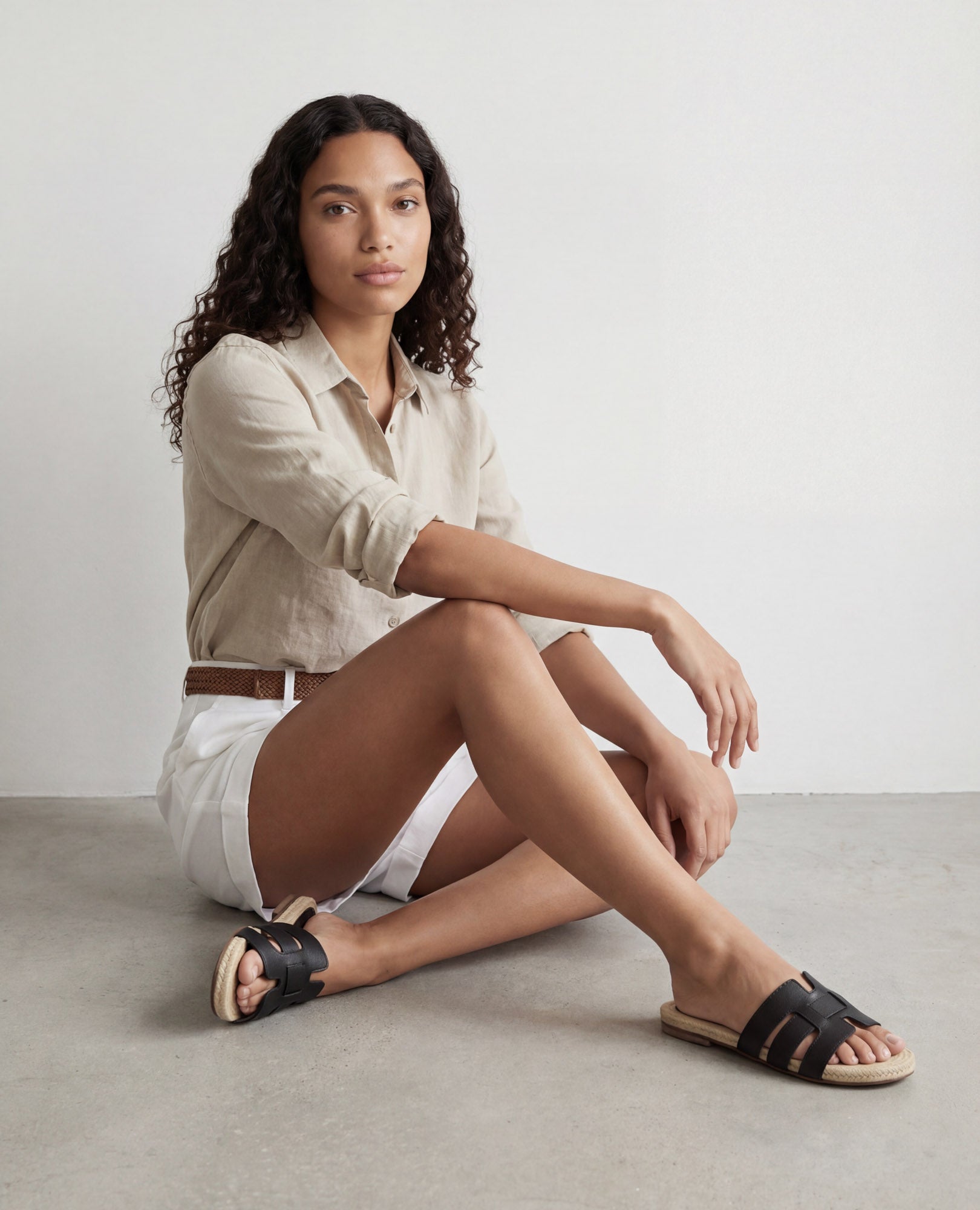 אישה עם שיער מתולתל לובשת חולצה בצבע בז', מכנסיים קצרים לבנים וכפכפים שטוחים של B.unique – תבל, יושבת על הרצפה.