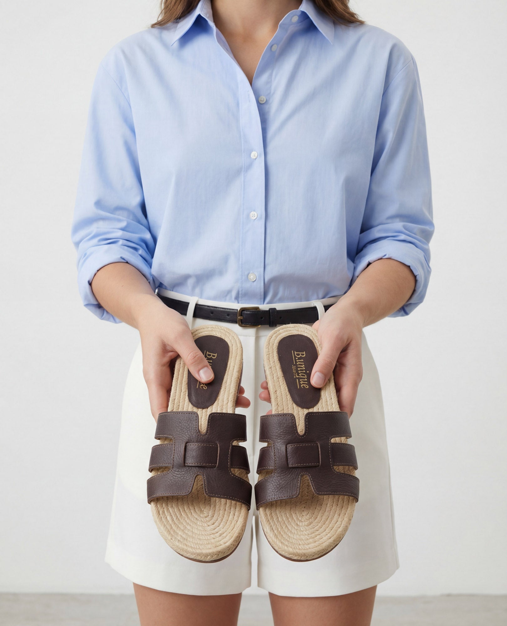 אדם לבוש בחולצה תכלת ובמכנסיים קצרים לבנים מחזיק כפכפים שטוחים בצבע חום של המותג B.unique.