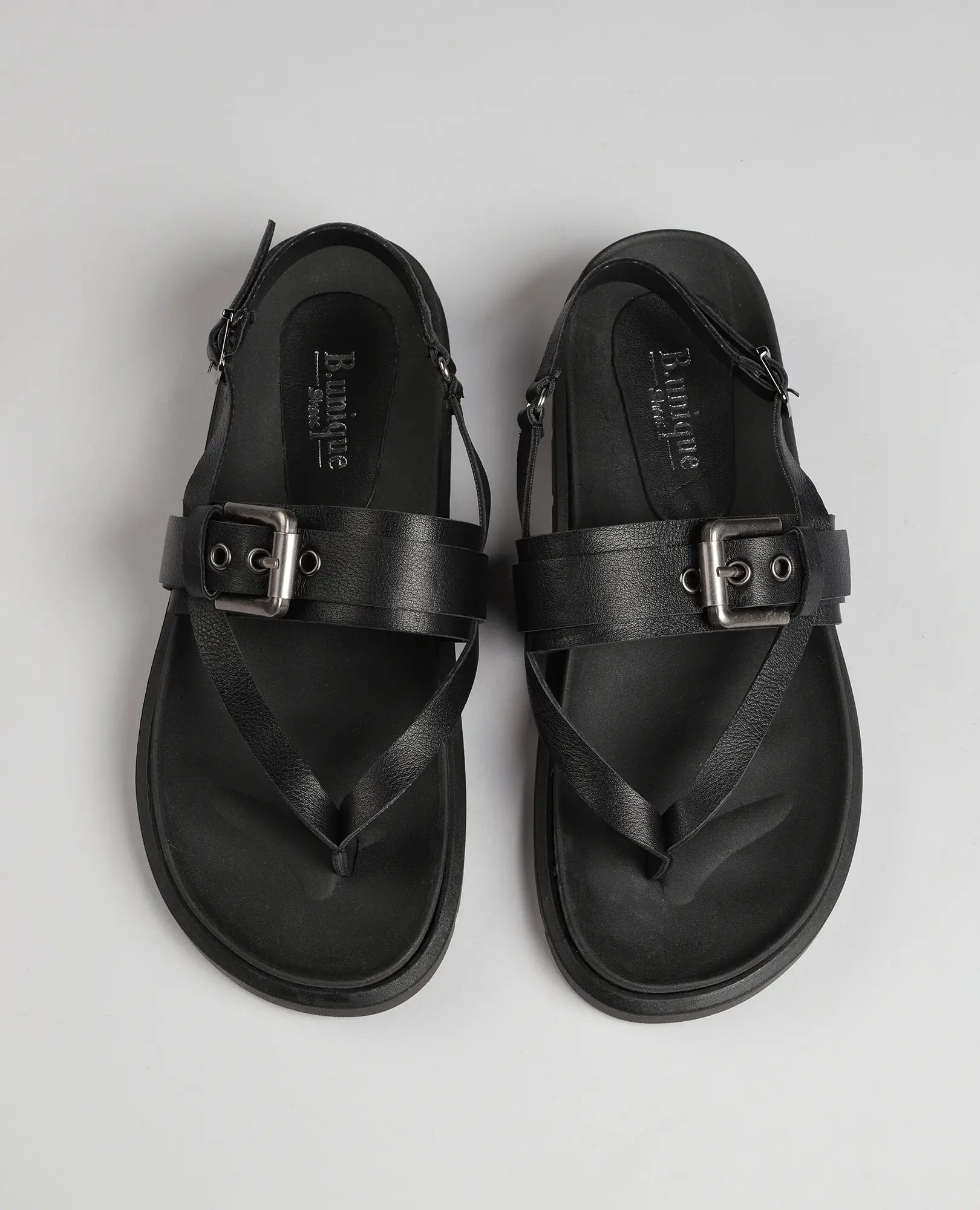 זוג סנדלי אצבע-פואט שחורים מבית B.unique עם אבזמים, רצועות קרסול ולולאות באצבעות, על רקע אפור בהיר.