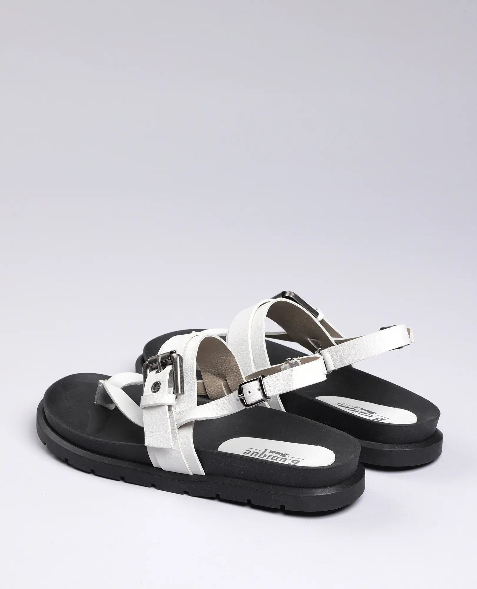 זוג סנדלי אצבע B.unique מדגם "פואט" בצבע לבן עם סוליות שחורות ואבזמים כסופים, על רקע אפור בהיר.