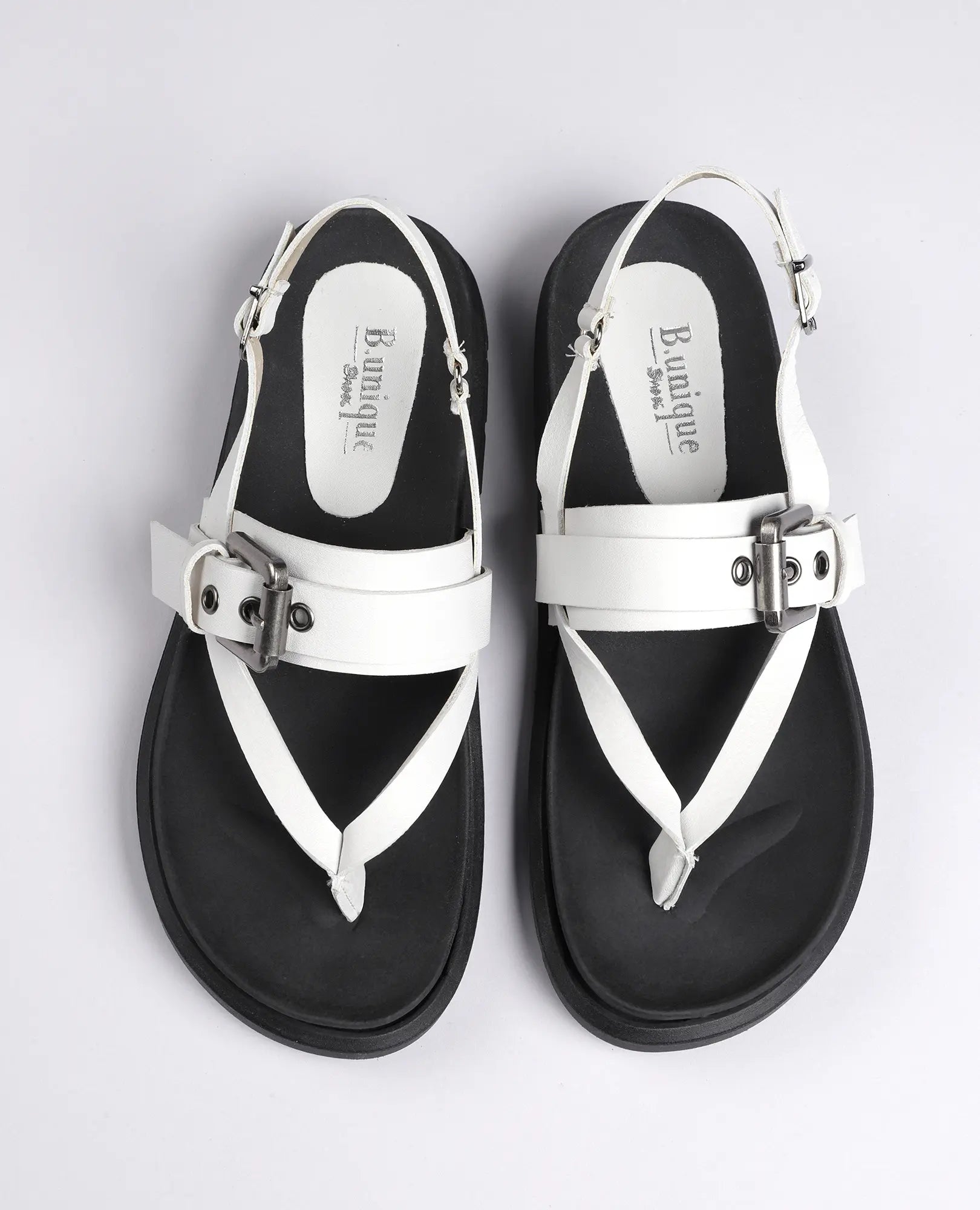 מבט מלמעלה על סנדלי האצבע "פואט" של B.unique, עם רצועות עור לבנות, אבזמים דקורטיביים וסוליות שחורות.