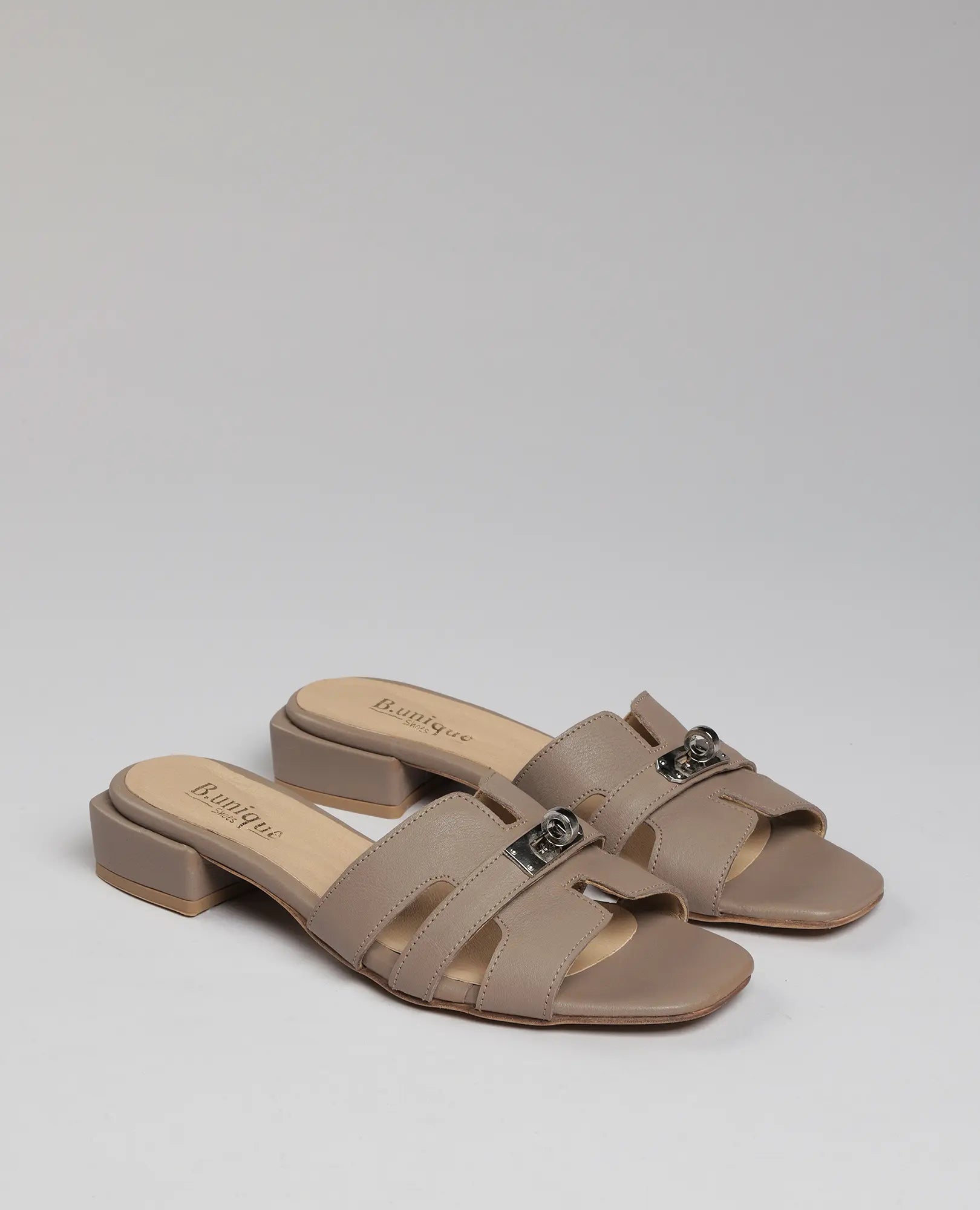 זוג כפכפי רצועות עם אבזם של B.unique - דגם "נעה" בצבע טאופ, עם רצועות עור רחבות ואבזמי מתכת על רקע בהיר.