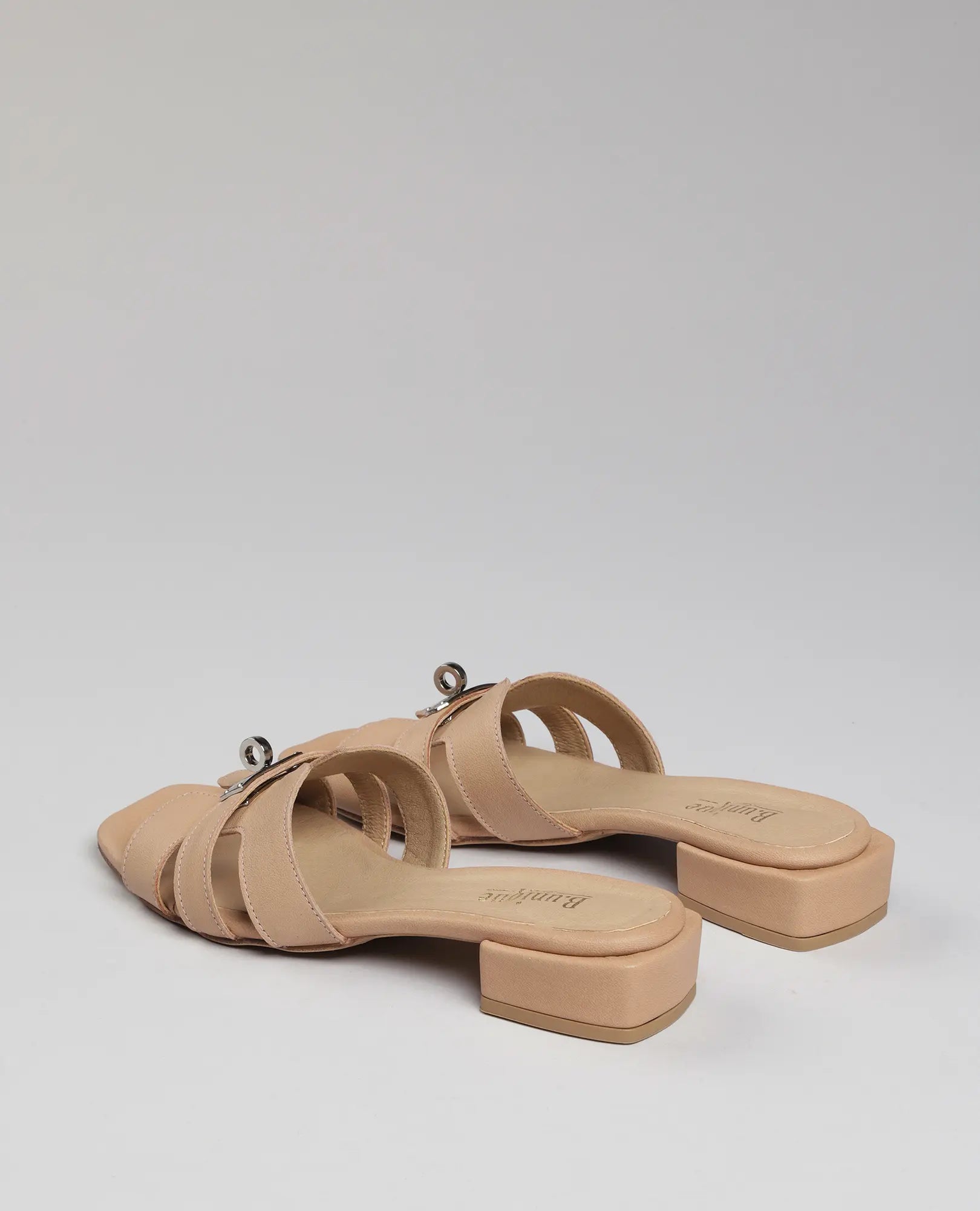 B.unique כפכפי רצועות עם אבזם - נעה: כפכפי עור בצבע בז', עם עקב, רצועות רחבות ואבזם כסוף על רקע לבן.