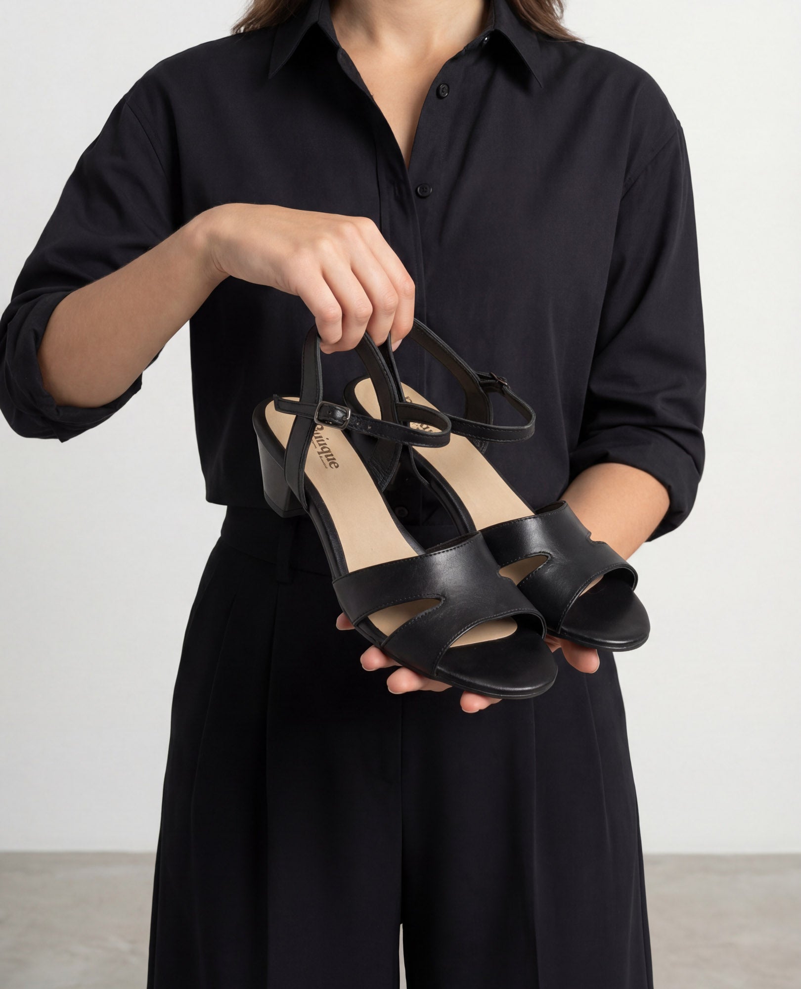 אישה לבושה בשחור מחזיקה בזוג סנדלי עקב "פלורנס שחור" מבית B.unique.