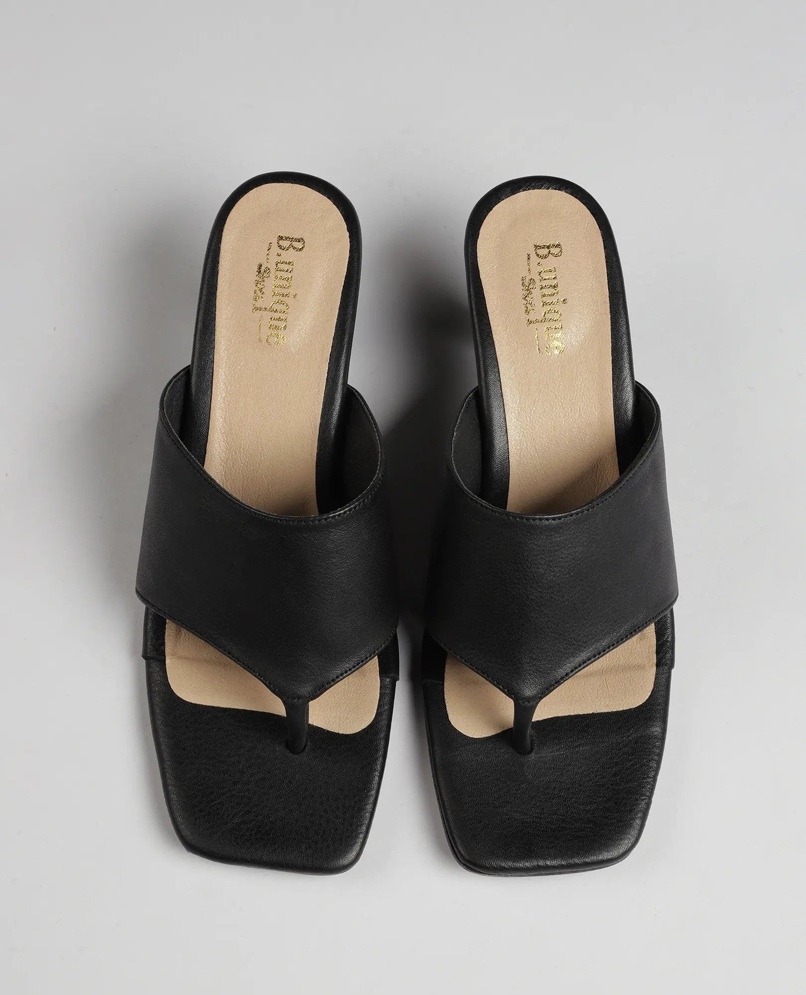 זוג כפכפי אצבע עם עקב של המותג B.unique, בצבע שחור עם רפידות בצבע בז' ועקבים בינוניים, על רקע אפור בהיר.