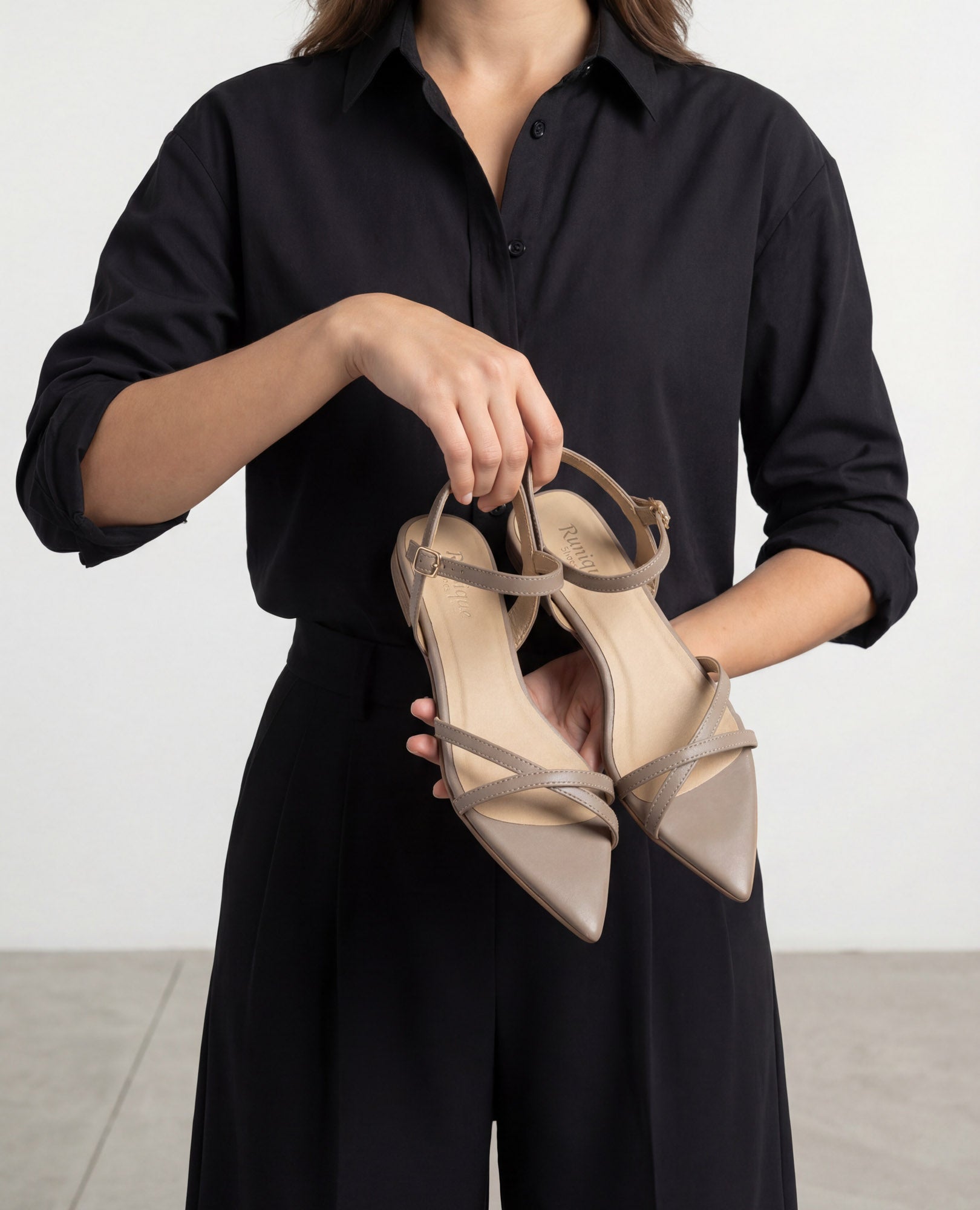 אדם לבוש בשחור מחזיק סנדלים שטוחות של המותג B.unique בצבע טאופ על רקע אחיד.