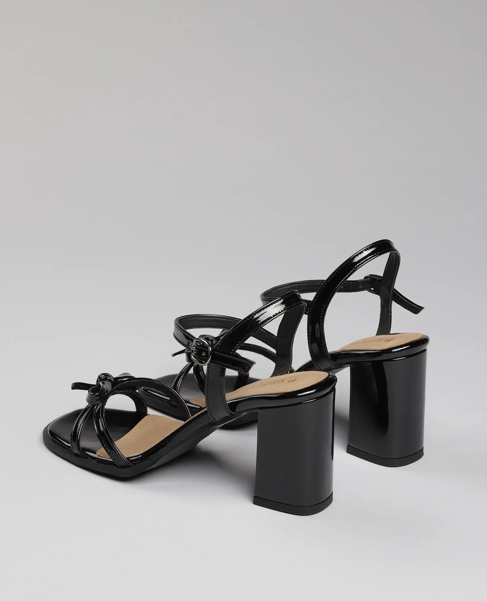 סנדלי עקב עם רצועת פפיון מבית B-Unique VEGAN בגימור דמוי עור בצבע שחור לכה, עם עקב בלוק ורצועות.