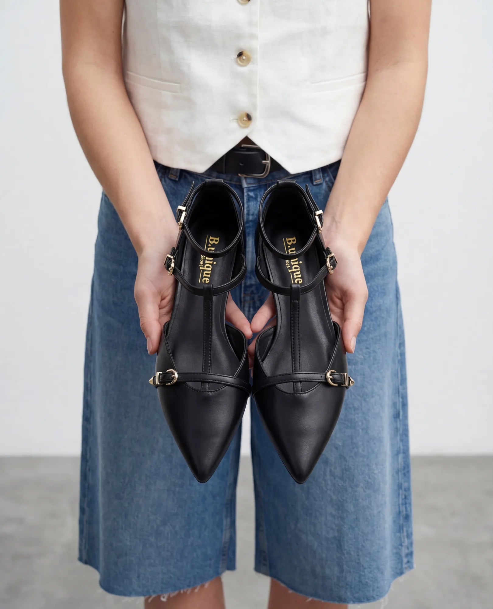 אישה לבושה באפודה לבנה וחצאית ג'ינס מחזיקה נעלי סירה שחורות - אושר מבית B-Unique VEGAN.