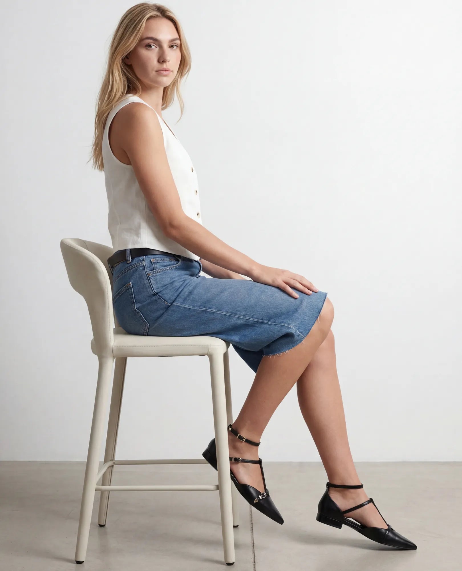 אישה לבושה בחולצה לבנה ללא שרוולים, חצאית ג'ינס ונעלי סירה של B-Unique VEGAN - יושבת בצד על כיסא גבוה, פונה למצלמה.