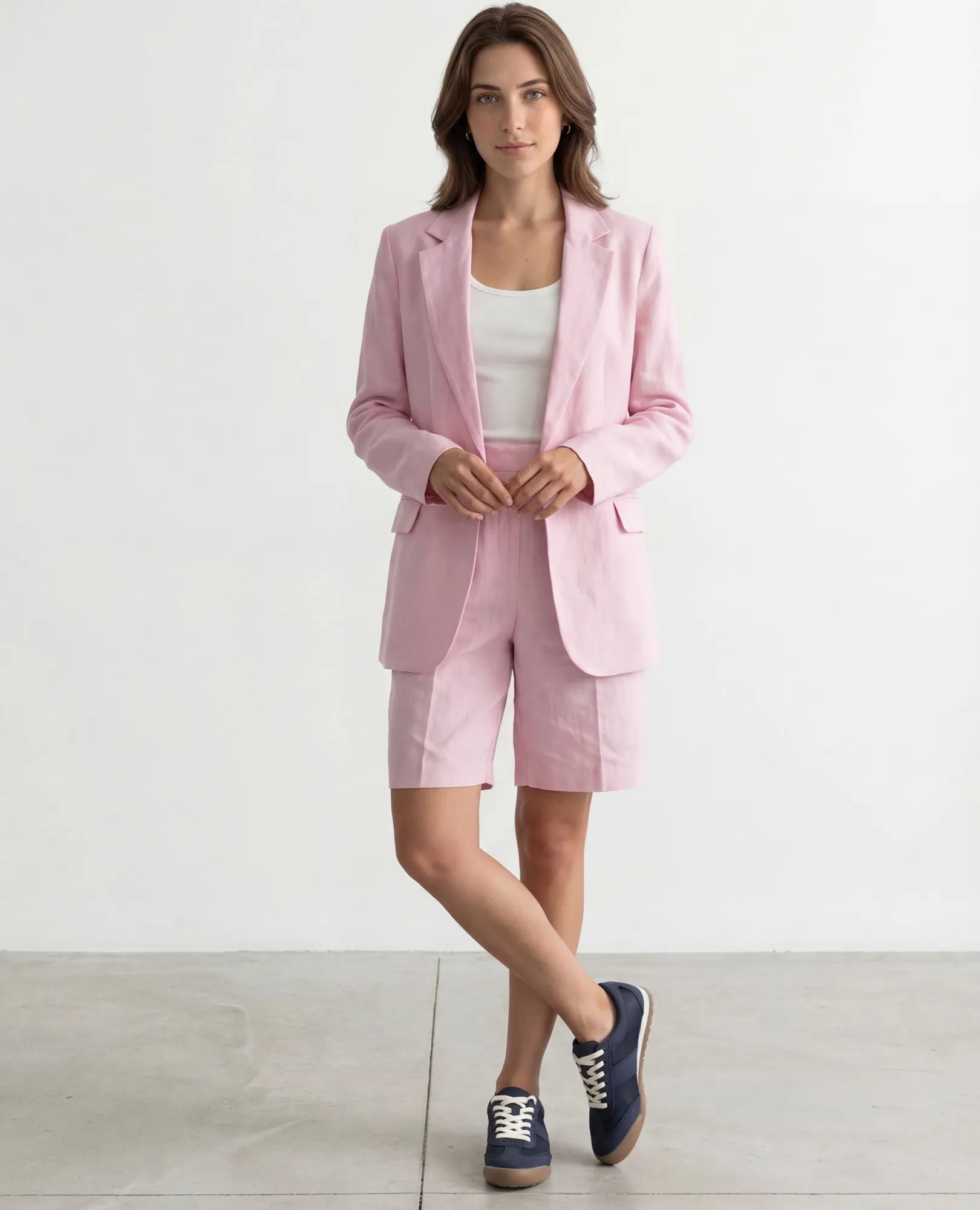 אישה לבושה בבלזר ורוד בהיר, מכנסיים קצרים, חולצה לבנה ונעלי סניקרס B-Unique VEGAN על רקע לבן חלק.