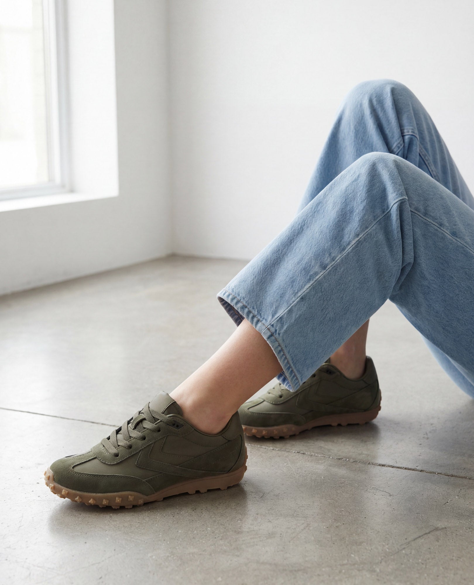 אדם לבוש בג'ינס תכלת וסניקרס מעור זמש של B.unique יושב על רצפת בטון מלוטשת ליד חלון.