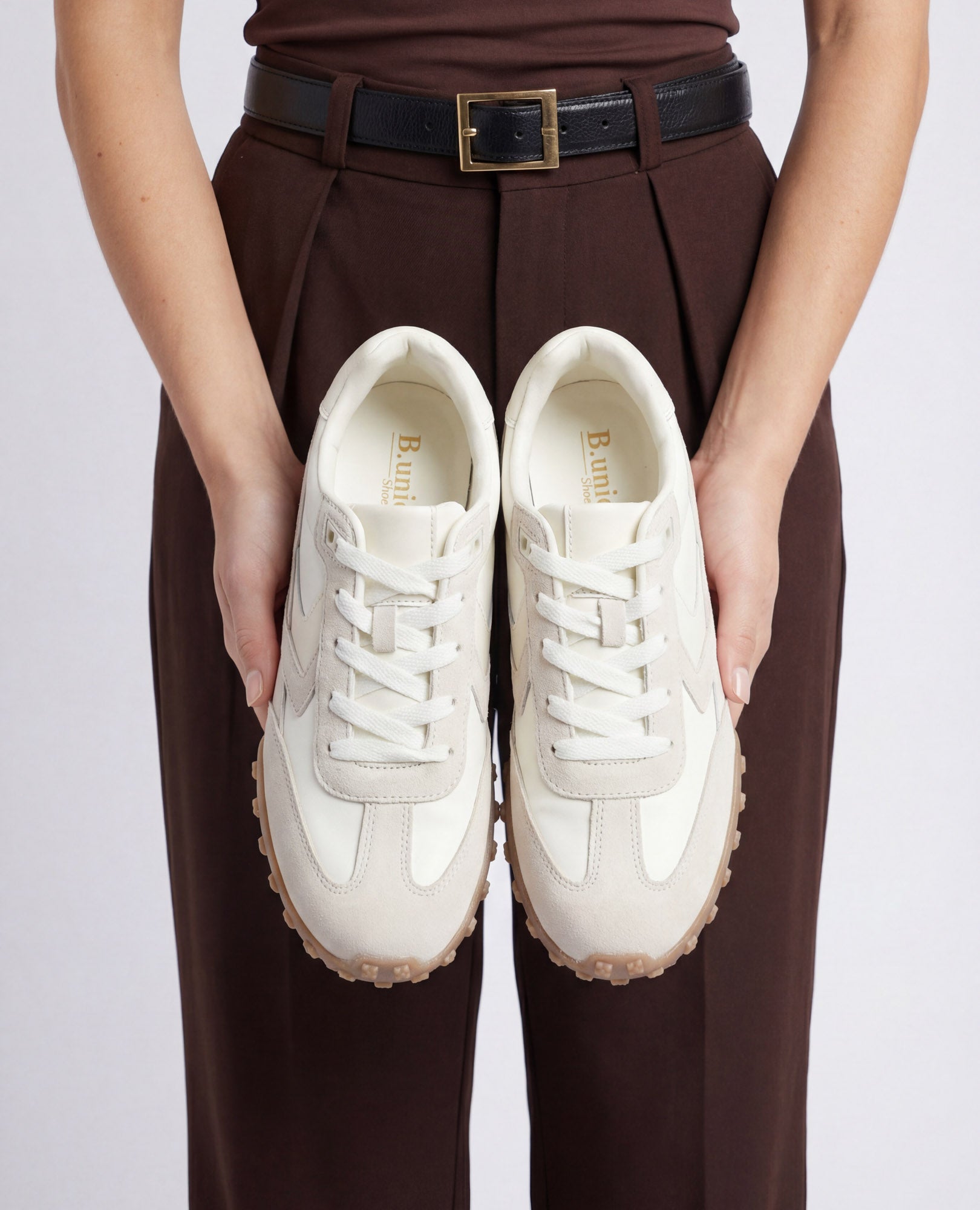 אדם במכנסיים חומים מחזיק זוג נעלי סניקרס מעור זמש-נטשה של B.unique עם סוליות בצבע בז', במבט מלמעלה.