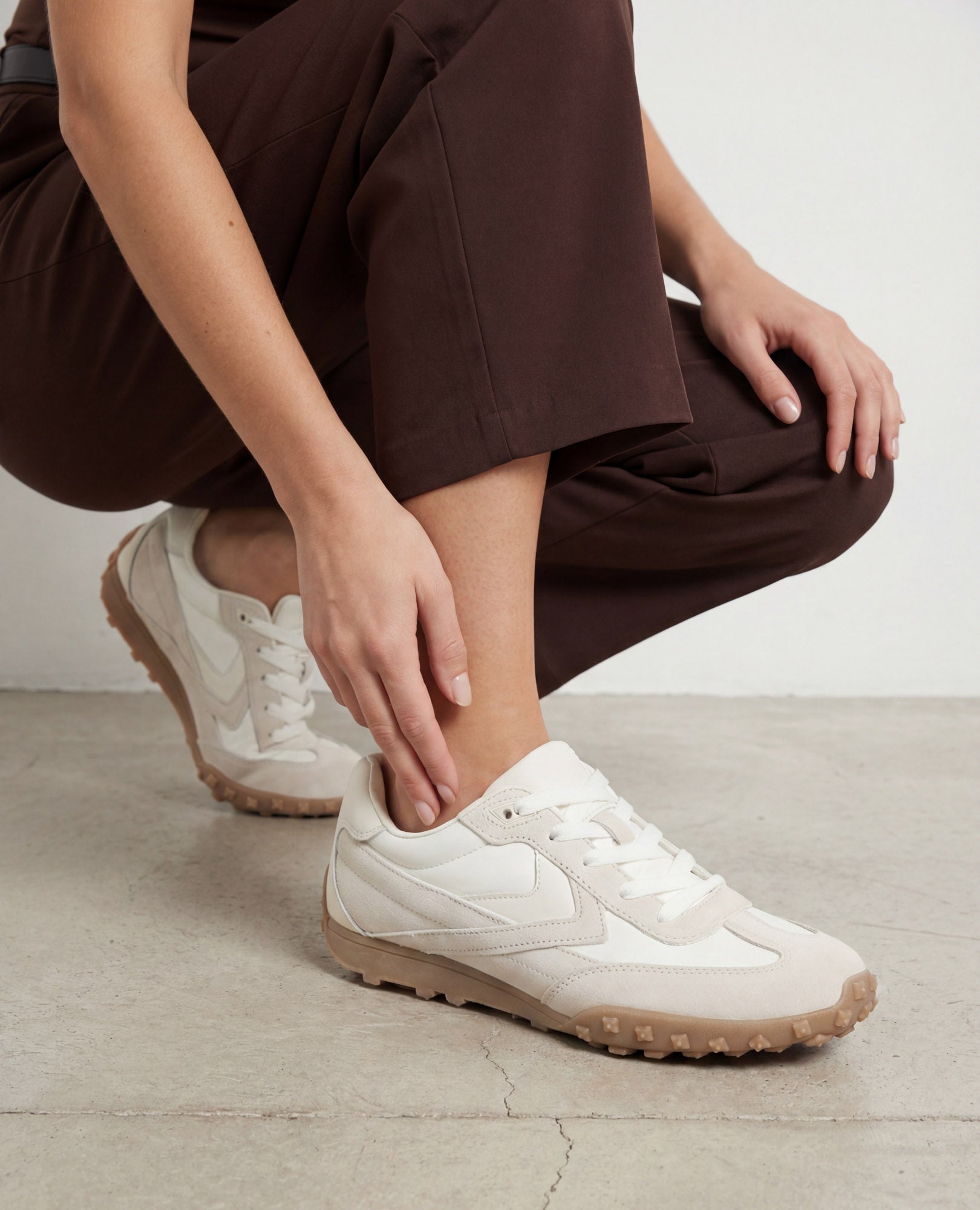 אדם במכנסיים חומים כורע על בטון, לובש נעלי סניקרס מעור זמש של B.unique בצבע לבן-שבור ובז' זמש.