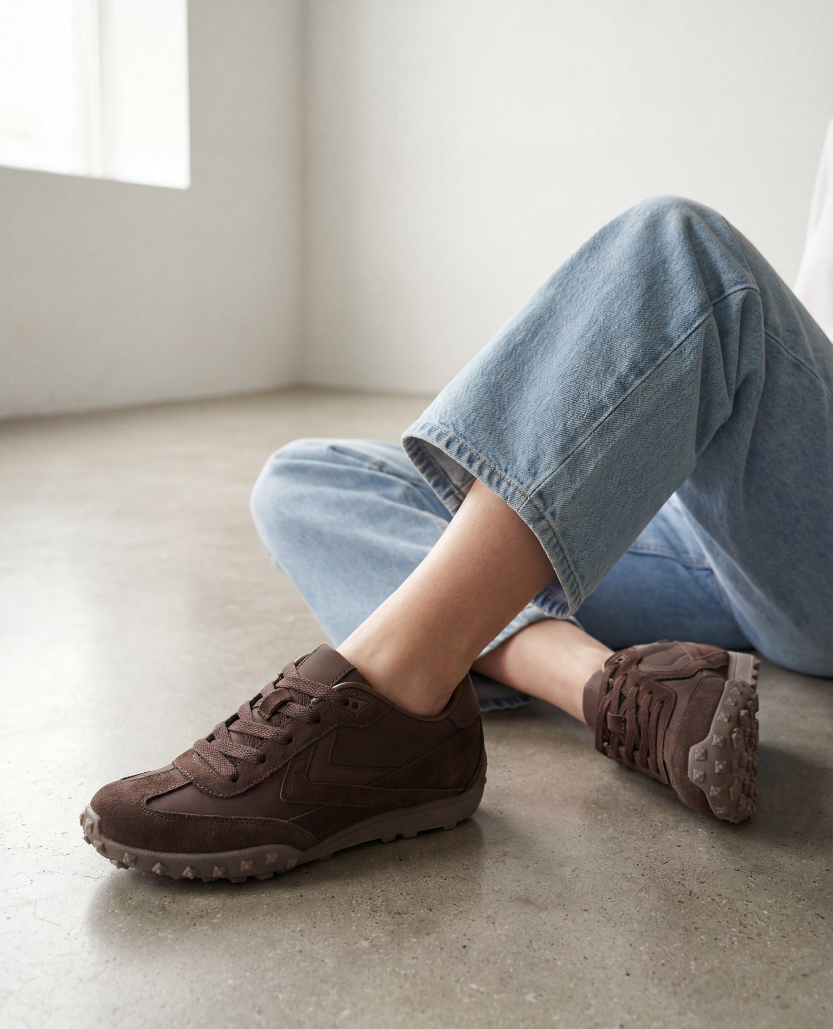 אדם לבוש בג'ינס תכלת וסניקרס מעור זמש- נטשה מבית B.unique יושב על רצפת בטון מלוטשת ליד חלון.