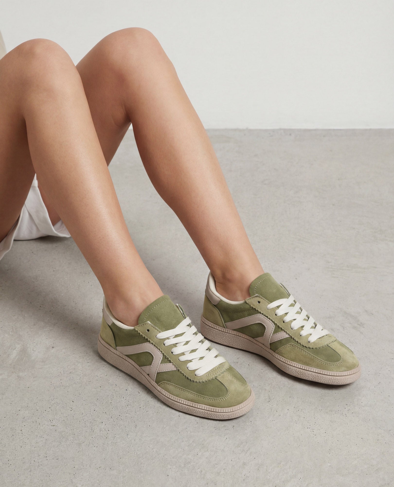 אדם יושב על רצפת בטון ולובש סניקרס דמוי זמש- נווה מבית B-Unique VEGAN בצבע ירוק זית ובז' עם שרוכים לבנים.