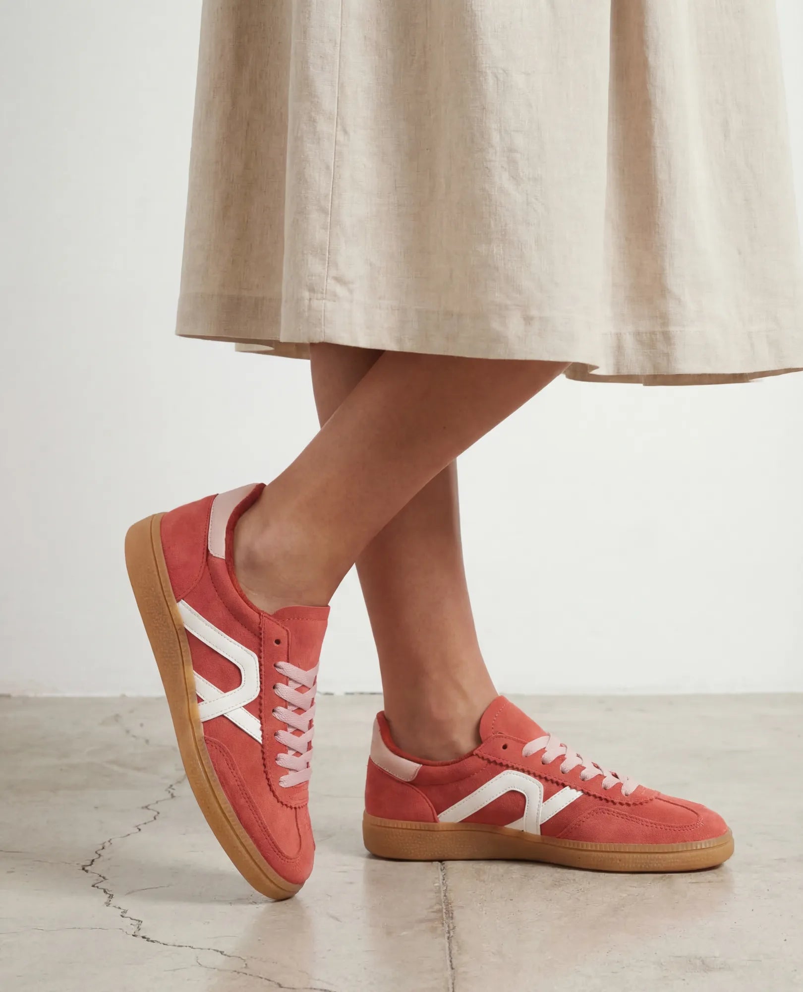 אישה בחצאית בצבע בז' נועלת נעלי ספורט B-Unique VEGAN דמוי זמש - נווה בצבע ורוד אלמוג עם סוליה לבנה וסוליה בצבע גומי על רצפת בטון.