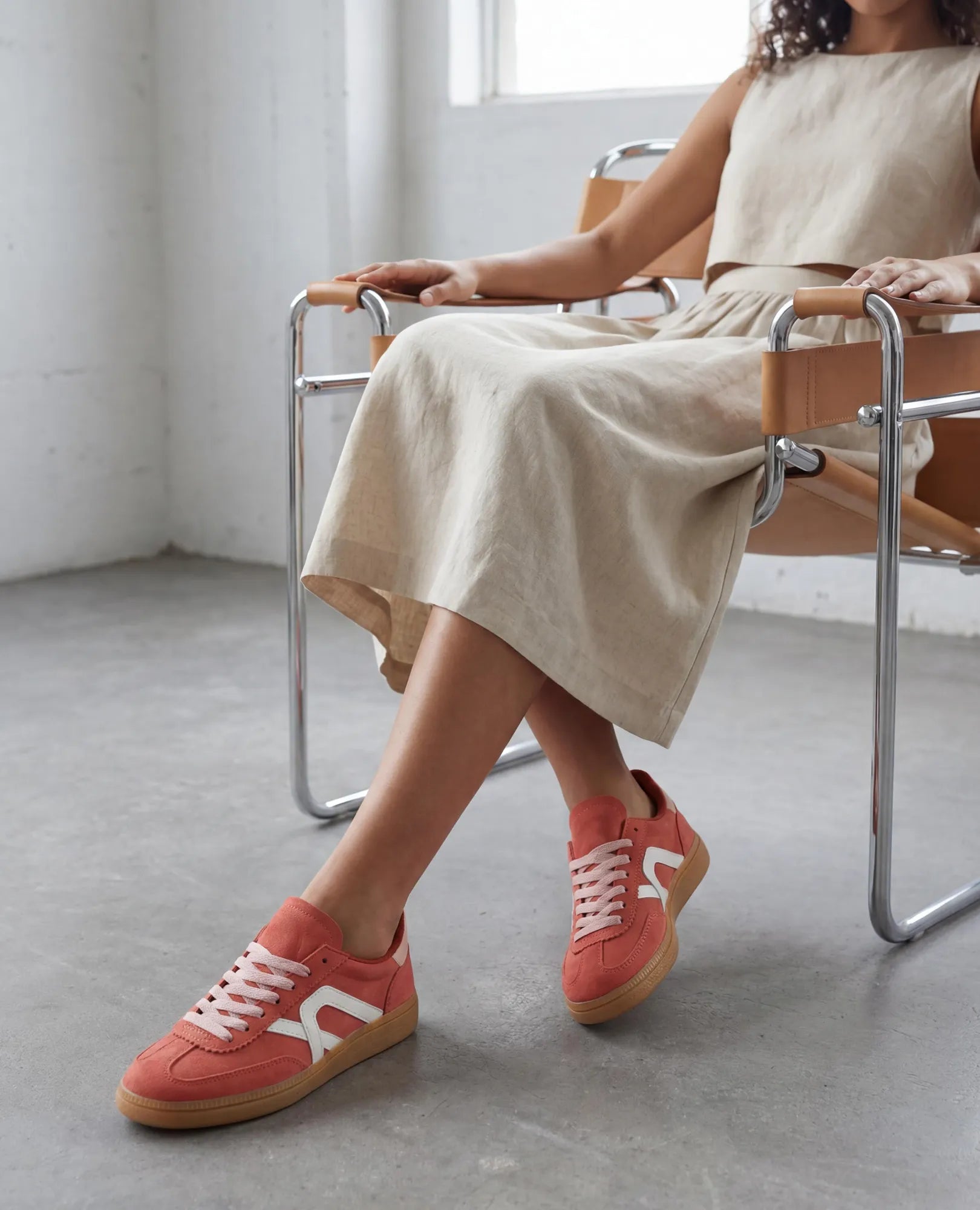 אישה לבושה בשמלה בצבע בז' יושבת על כיסא מודרני, נועלת נעלי ספורט B-Unique VEGAN דמוי זמש - נווה עם סוליות לבנות וצבע גומי.