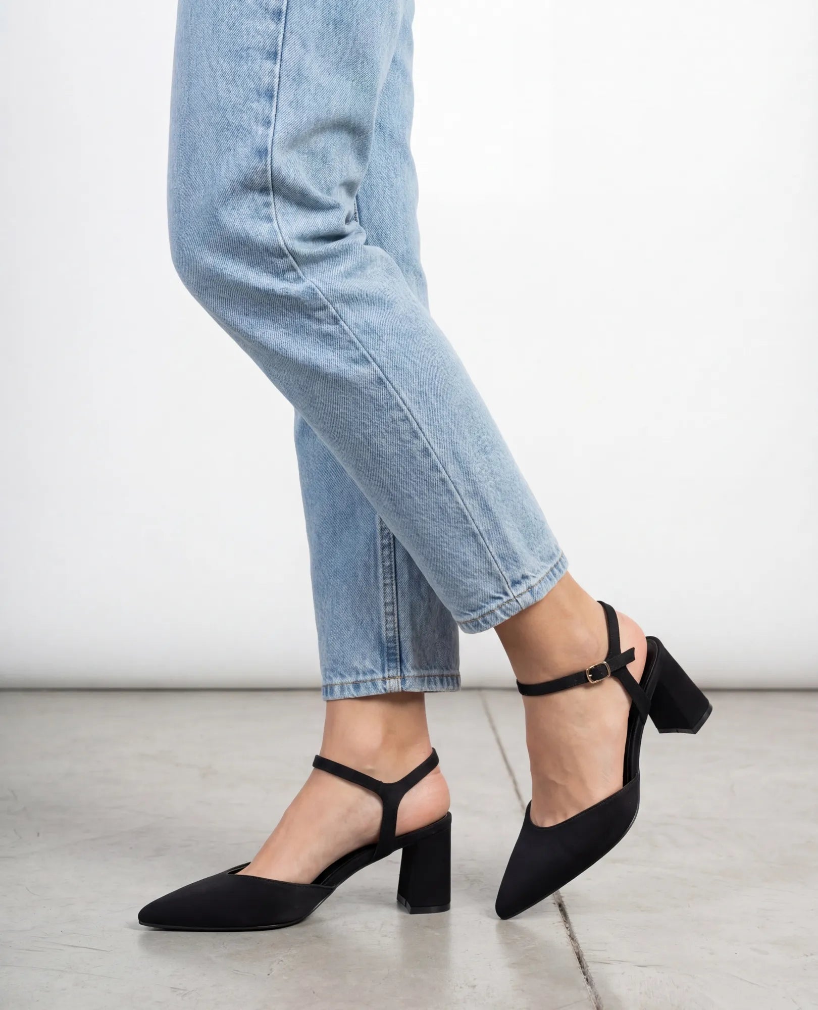 אישה לבושה בג'ינס תכלת ונעלי עקב – "ארין שחור" מבית B-Unique VEGAN, עומדת על רצפת בטון.