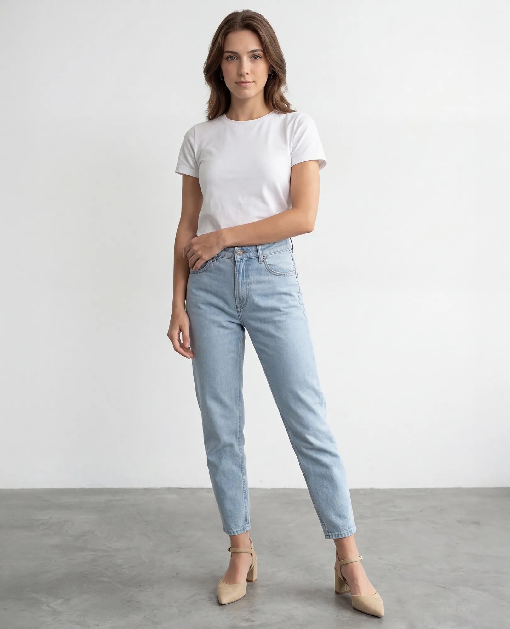 אישה לבושה בחולצת טי לבנה, ג'ינס תכלת ונעלי עקב בצבע בז' – דגם "ארין" מבית B-Unique VEGAN – על רקע לבן חלק.