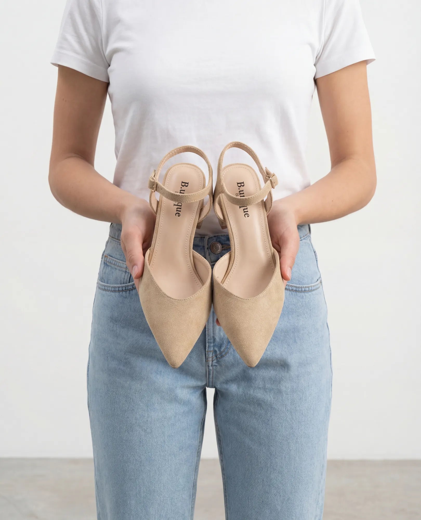 אישה בחולצת טי לבנה ובג'ינס כחול, אוחזת בנעלי עקב - דגם "ארין" של המותג B-Unique VEGAN בצבע קאמל.