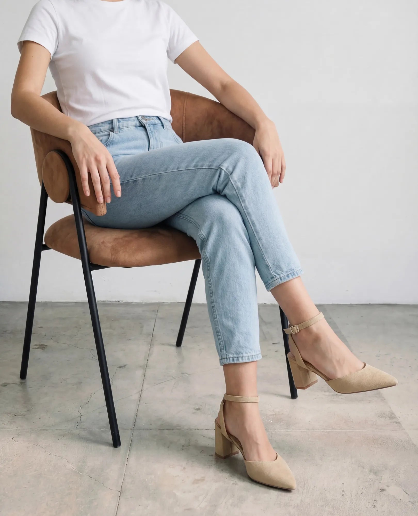 אישה יושבת על כיסא, לבושה בחולצת טי לבנה, ג'ינס תכלת ונעלי עקב-ארין של B-Unique VEGAN, כשהיא יושבת ברגליים משוכלות.