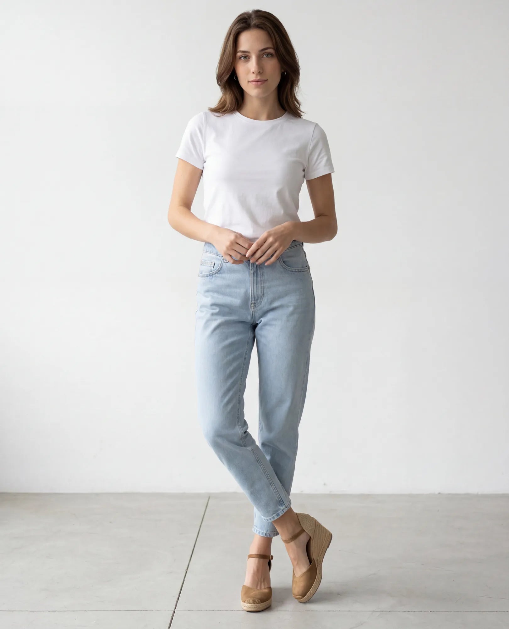 אישה לבושה בחולצת טי לבנה, ג'ינס כחול ונעלי פלטפורמה של B-Unique VEGAN – כרם עם רצועת קרסול – עומדת בחדר מואר ומינימליסטי.