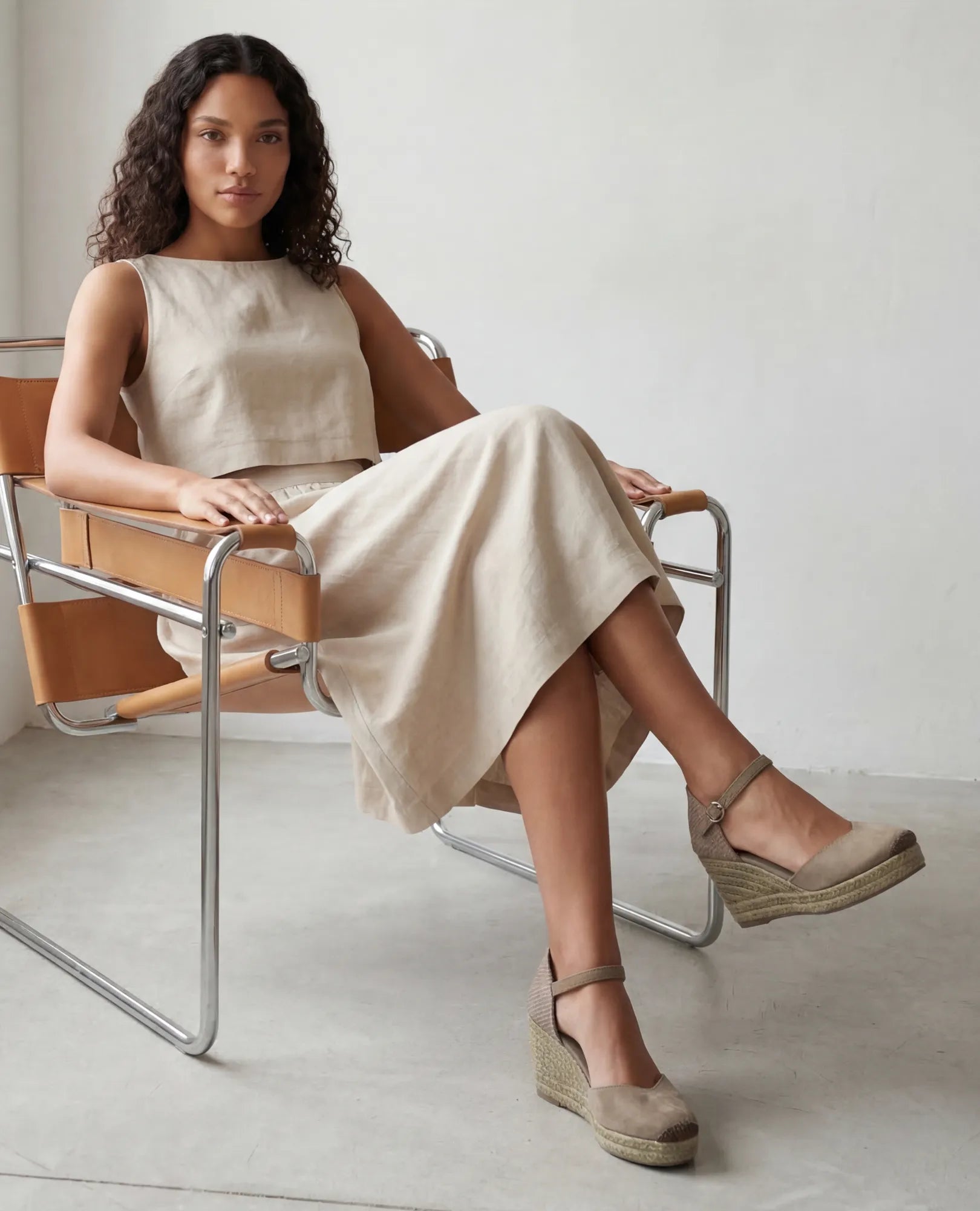 אישה לבושה בשמלה בצבע בז' ללא שרוולים ונעלי פלטפורמה של B-Unique VEGAN בצבע קרם עם רצועת קרסול יושבת על כיסא מודרני.