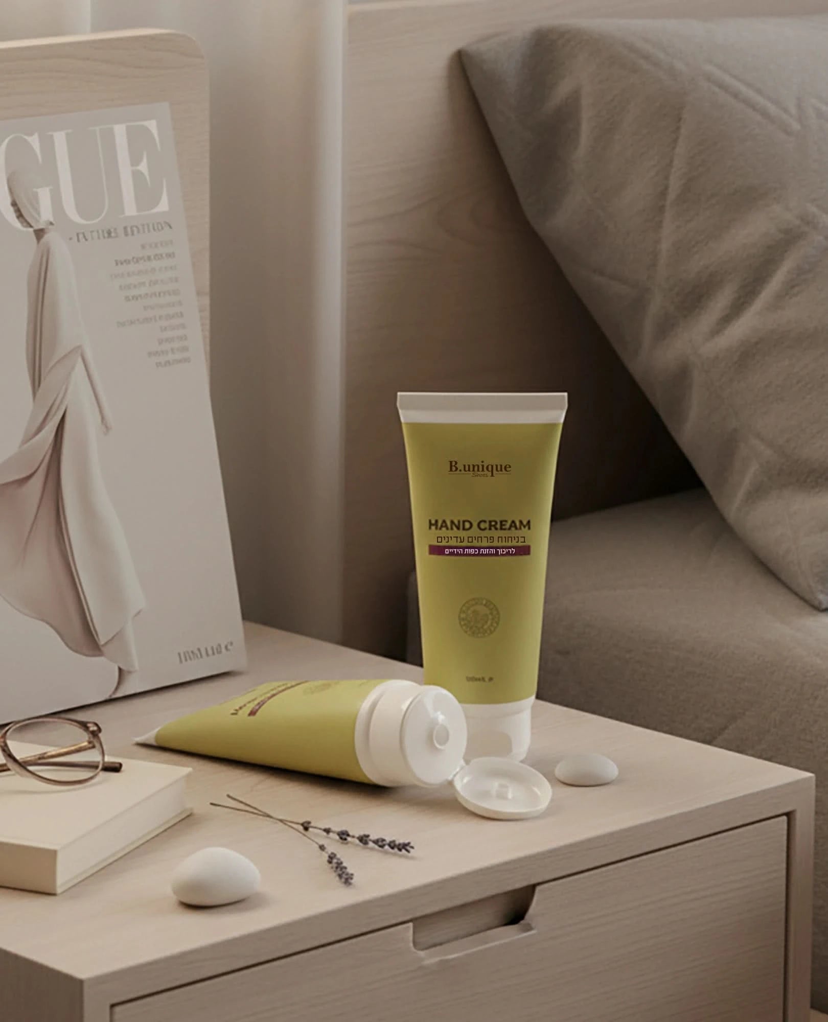 שני שפופרות של קרם ידיים בניחוח פרחים עדין, מגזין ומשקפיים מונחים על שולחן ליד מיטה אפורה.