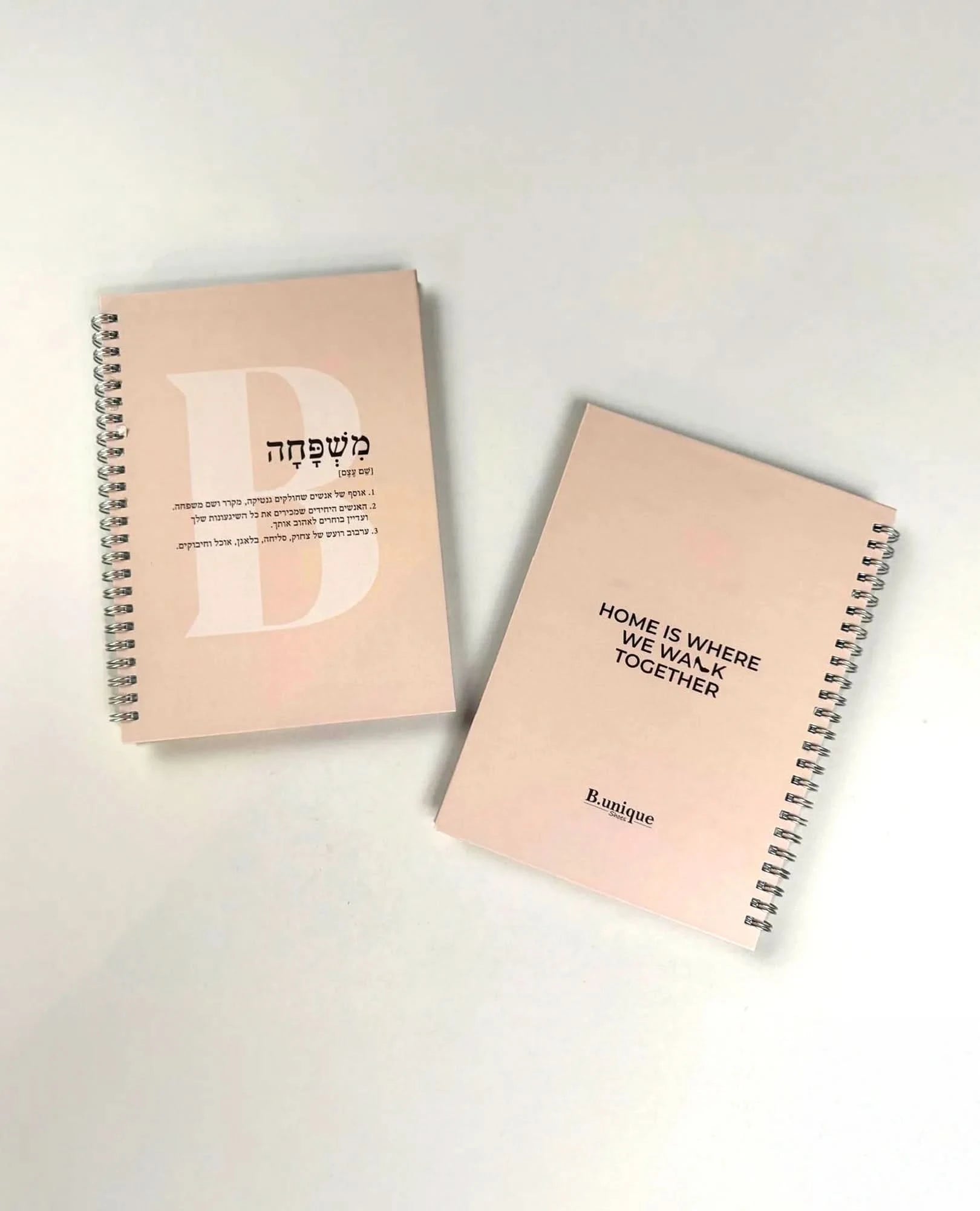 Two pink "מחברת שורות ליום המשפחה" notebooks rest on a white surface—one with Hebrew text and a bold B, the other with "HOME IS WHERE WE WALK TOGETHER" and "B-unique," inspired by קרם גוף scents and soothing שמנים אתריים.