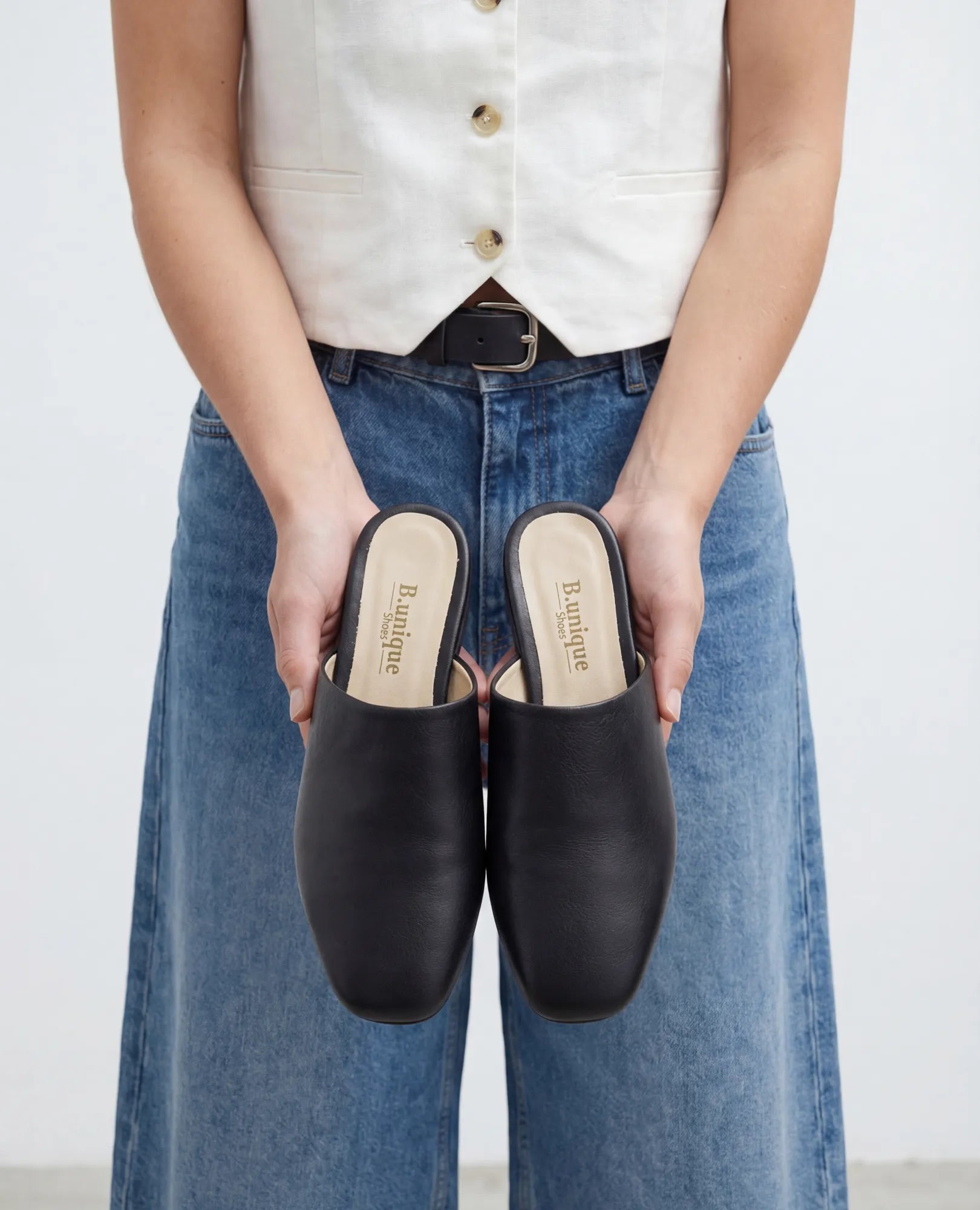 אדם לבוש באפוד לבן ובג'ינס כחול מחזיק כפכפים שטוחים של B.unique – רוקס – מול המצלמה.