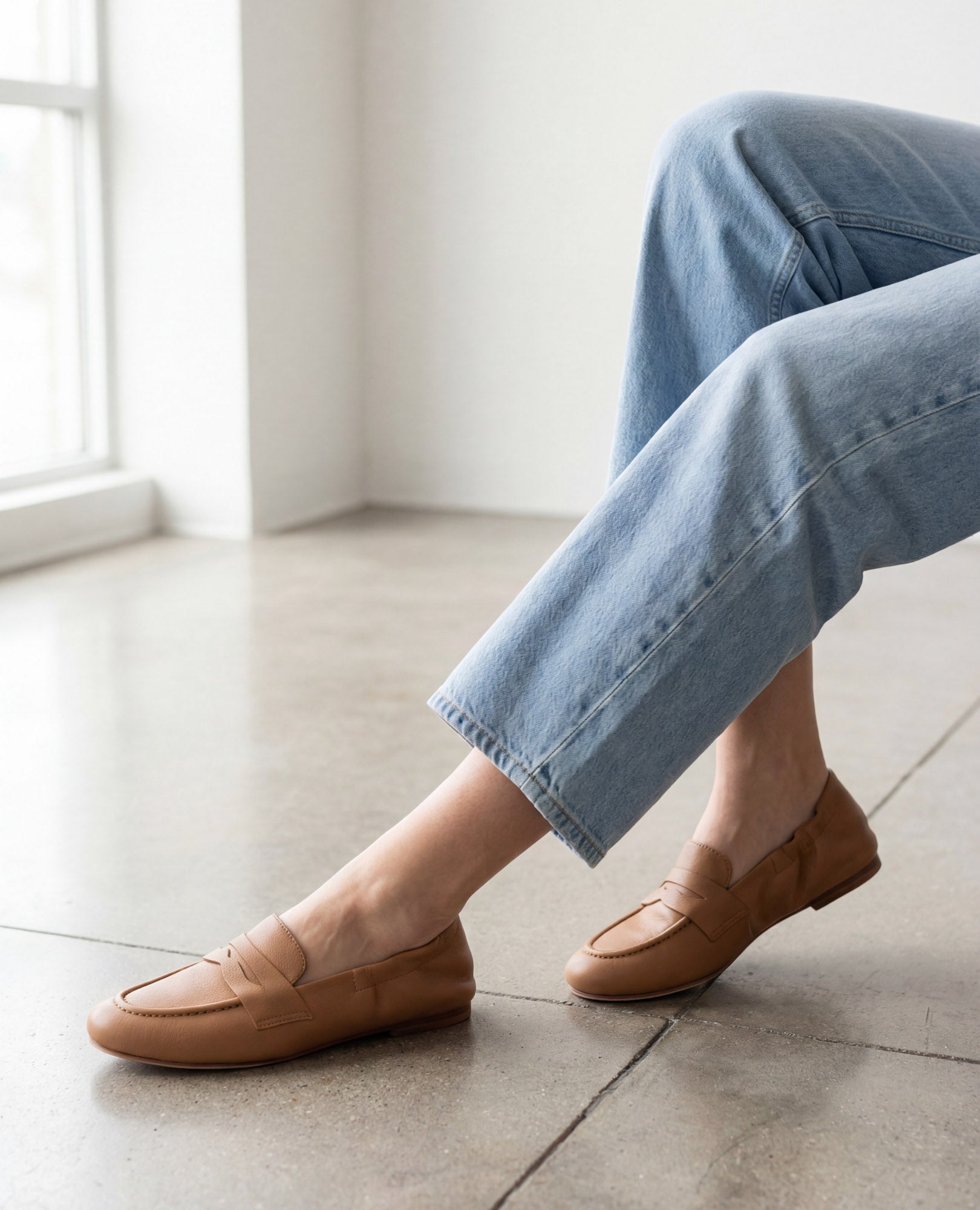 אדם לבוש בג'ינס תכלת ונעלי מוקסין-נוריאל של B.unique יושב בתוך הבית על רצפה מלוטשת ליד חלון.