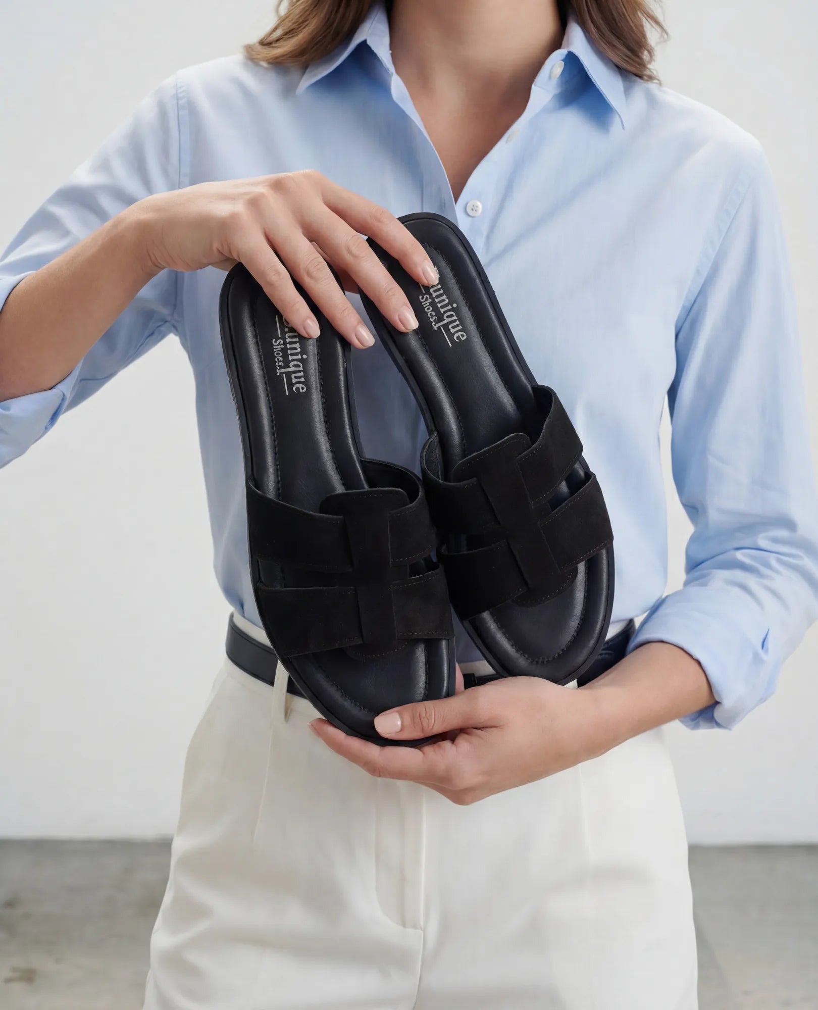 אדם לבוש בחולצה תכלת מחזיק כפכפים שטוחים – מורגן שחור של B.unique.