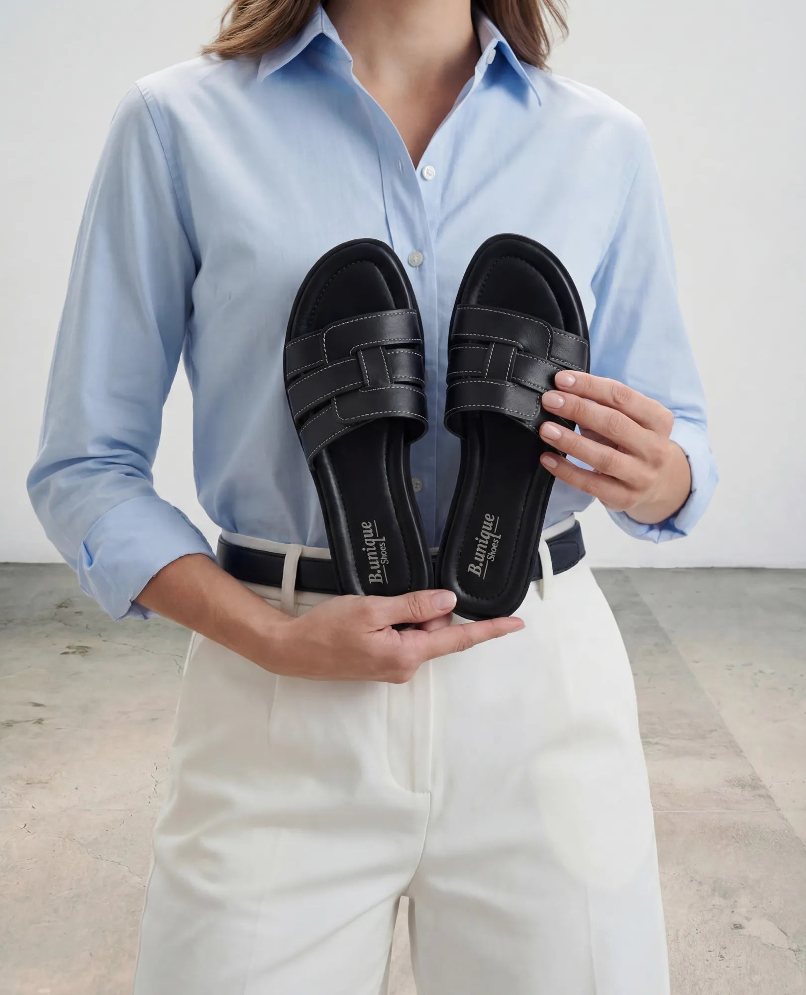 אישה לבושה בחולצה כחולה ומכנסיים לבנים מחזיקה את כפכפי הרצועות "B.unique" - סקאיי מול פלג גופה העליון.