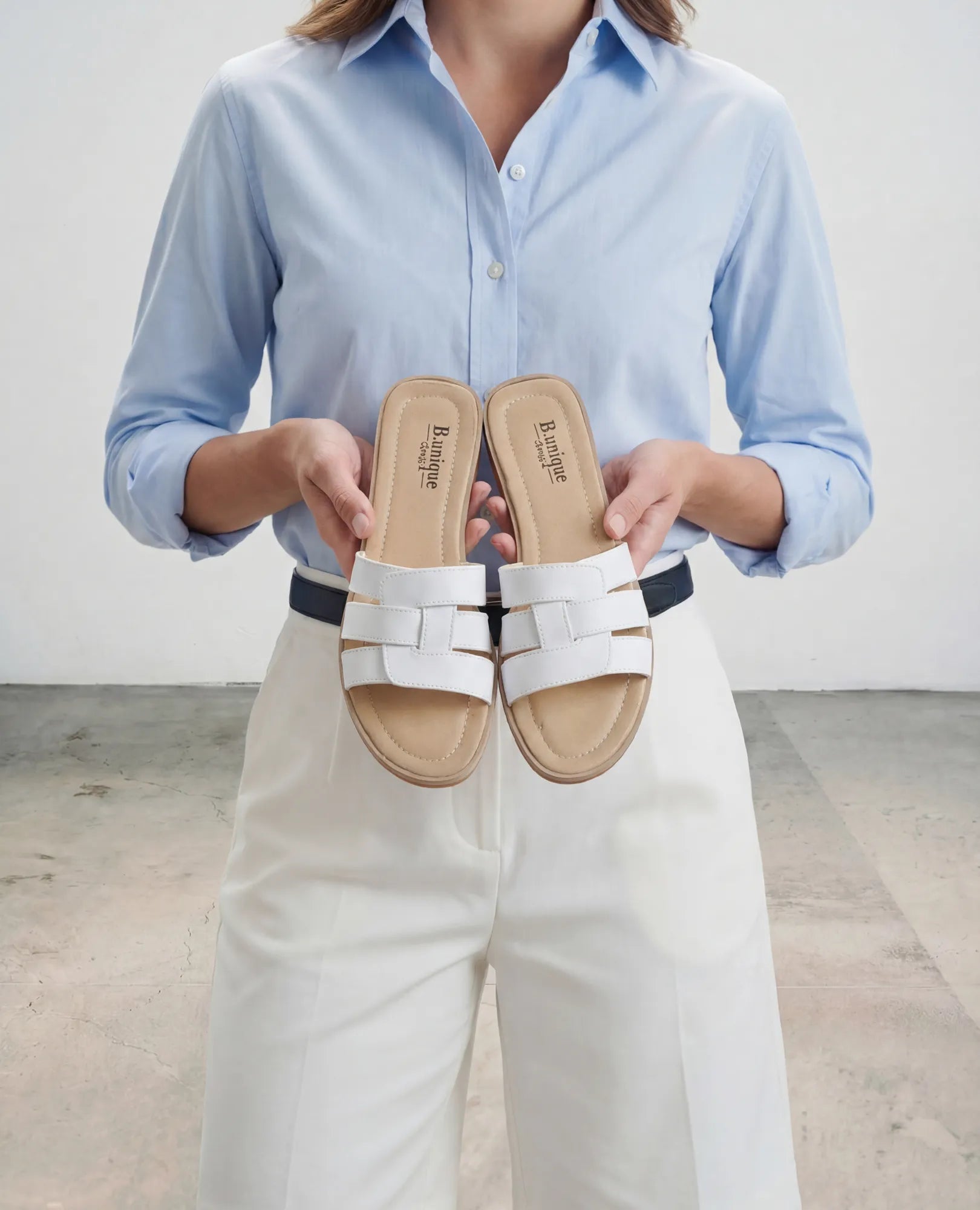 אישה בחולצה תכלת ומכנסיים לבנים, נועלת כפכפי רצועות - סקאיי מבית B.unique, עם רצועות עור וסוליות בצבע חום בהיר.