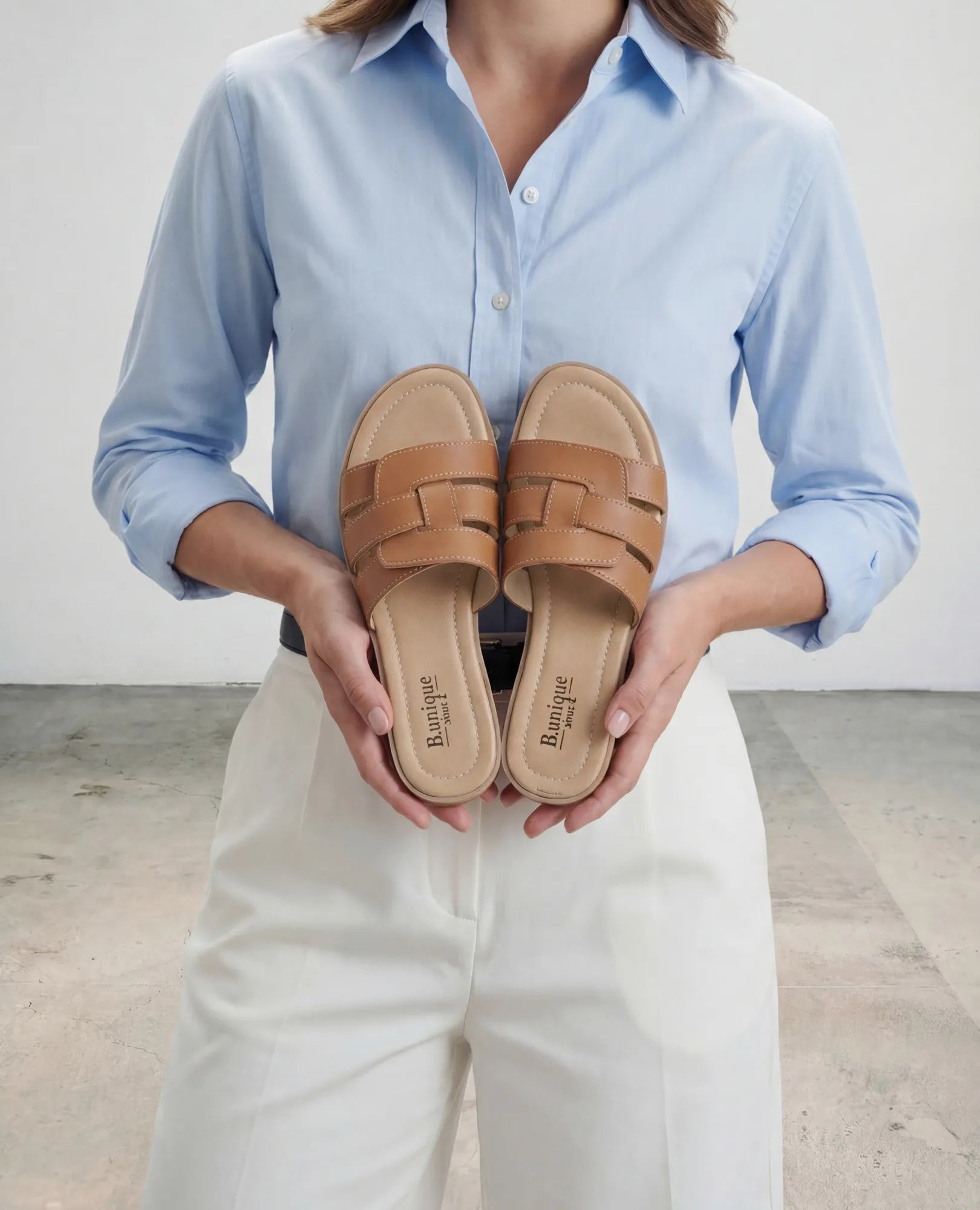 אישה לבושה בחולצה תכלת מחזיקה בידיה כפכפי רצועות - סקאיי מבית B.unique בצבע חום בהיר עם רצועות עור בחזית.