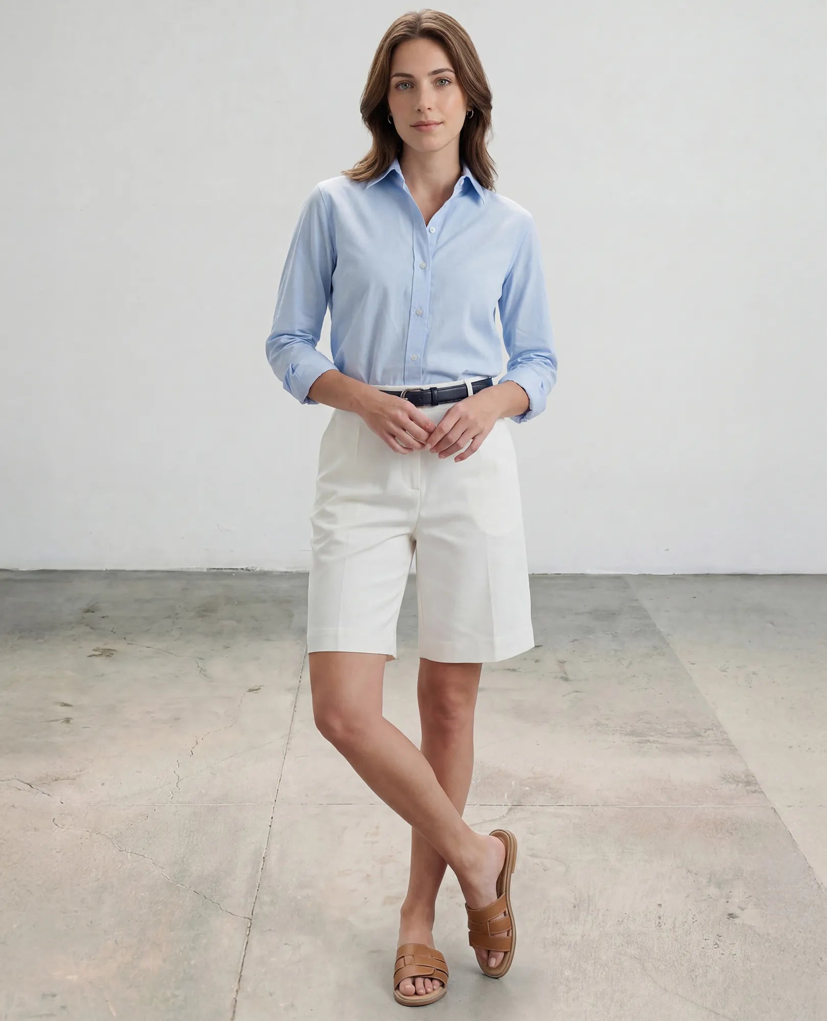 אישה לבושה בחולצה תכלת, מכנסיים קצרים לבנים וכפכפי רצועות של B.unique - סקאיי עומדת בחדר מינימליסטי.