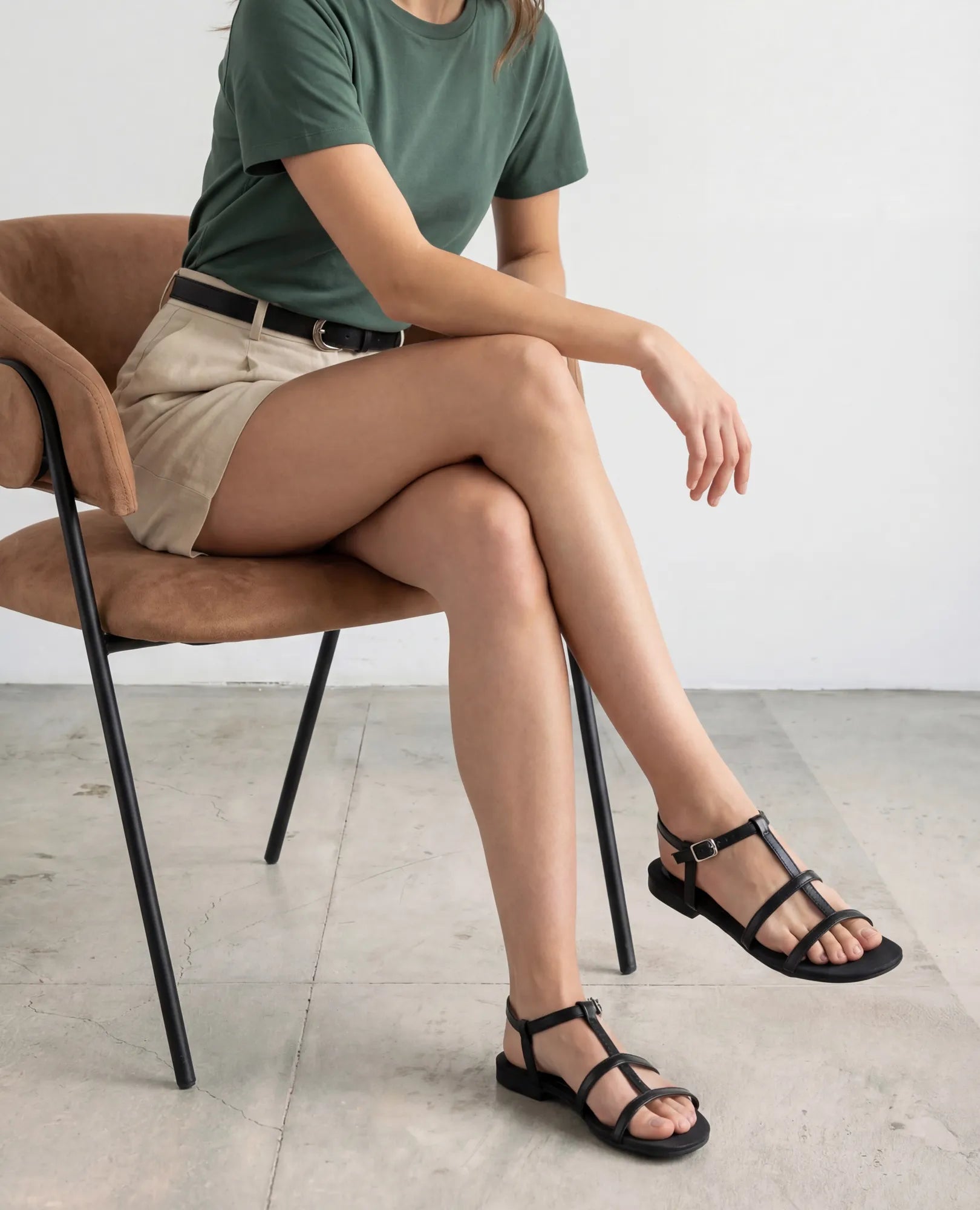 אישה לבושה בחולצת טי ירוקה ובמכנסיים קצרים בצבע בז' יושבת על רצפת בטון, לרגליה נעלי גלדיאטור של המותג B.unique מדגם "קיילי".